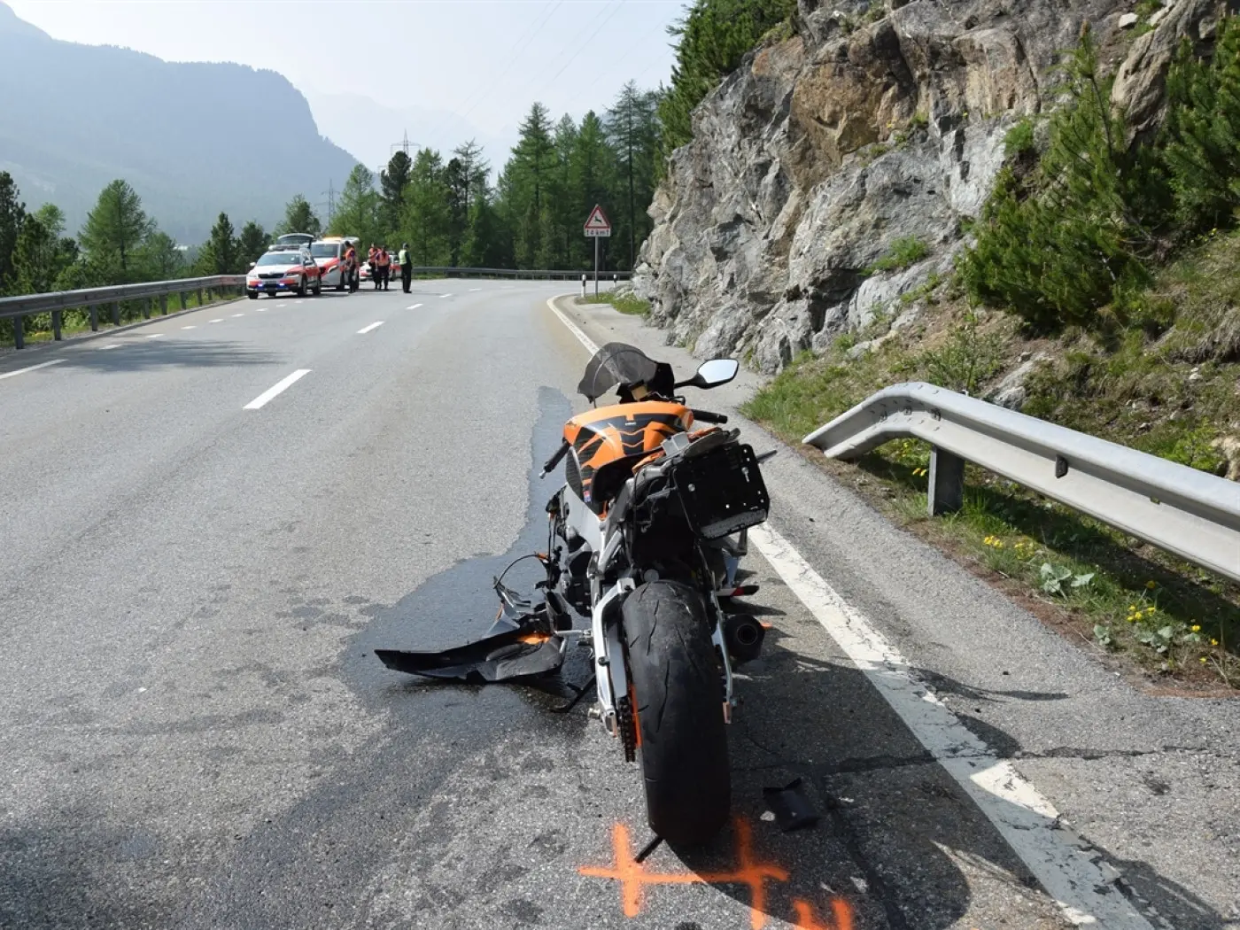 Una moto danneggiata sul lato di una strada montana. Ci sono marcature arancioni a terra e alcuni soccorritori visibili in lontananza. Il paesaggio circostante è montuoso con alberi.