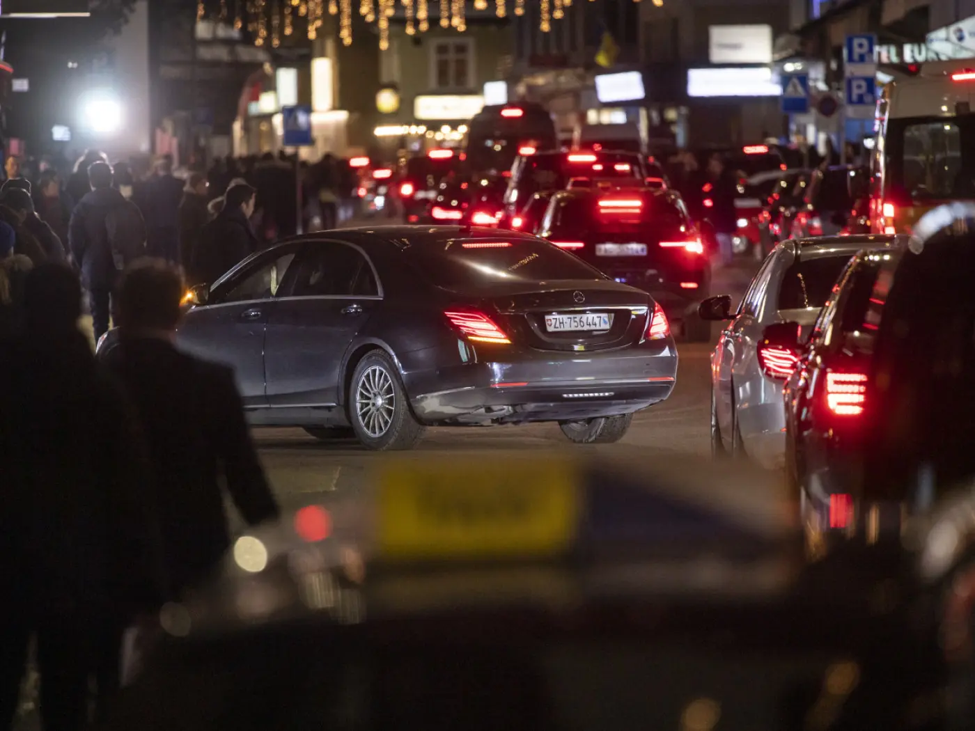 Una strada affollata di notte con auto in movimento e persone che camminano. Un'auto nera si sta immettendo nella carreggiata mentre altre auto sono parcheggiate lungo il marciapiede. Luci decorative illuminano l'ambiente.