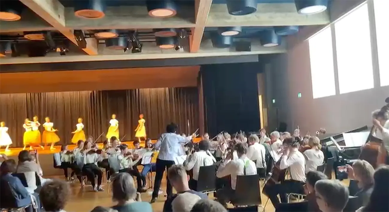 Un'orchestra si esibisce su un palco mentre ballerini si esibiscono in un balletto. Il pubblico è seduto in sala, affascinato dalla performance.