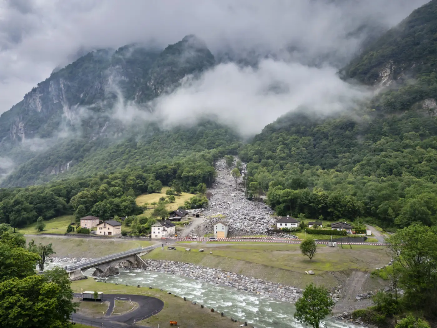 Uno scenario montano con nuvole basse che coprono le vette. Una frana di rocce scende dalla montagna verso un villaggio. Case e una strada si trovano ai piedi della montagna, mentre un fiume scorre attraverso il paesaggio.