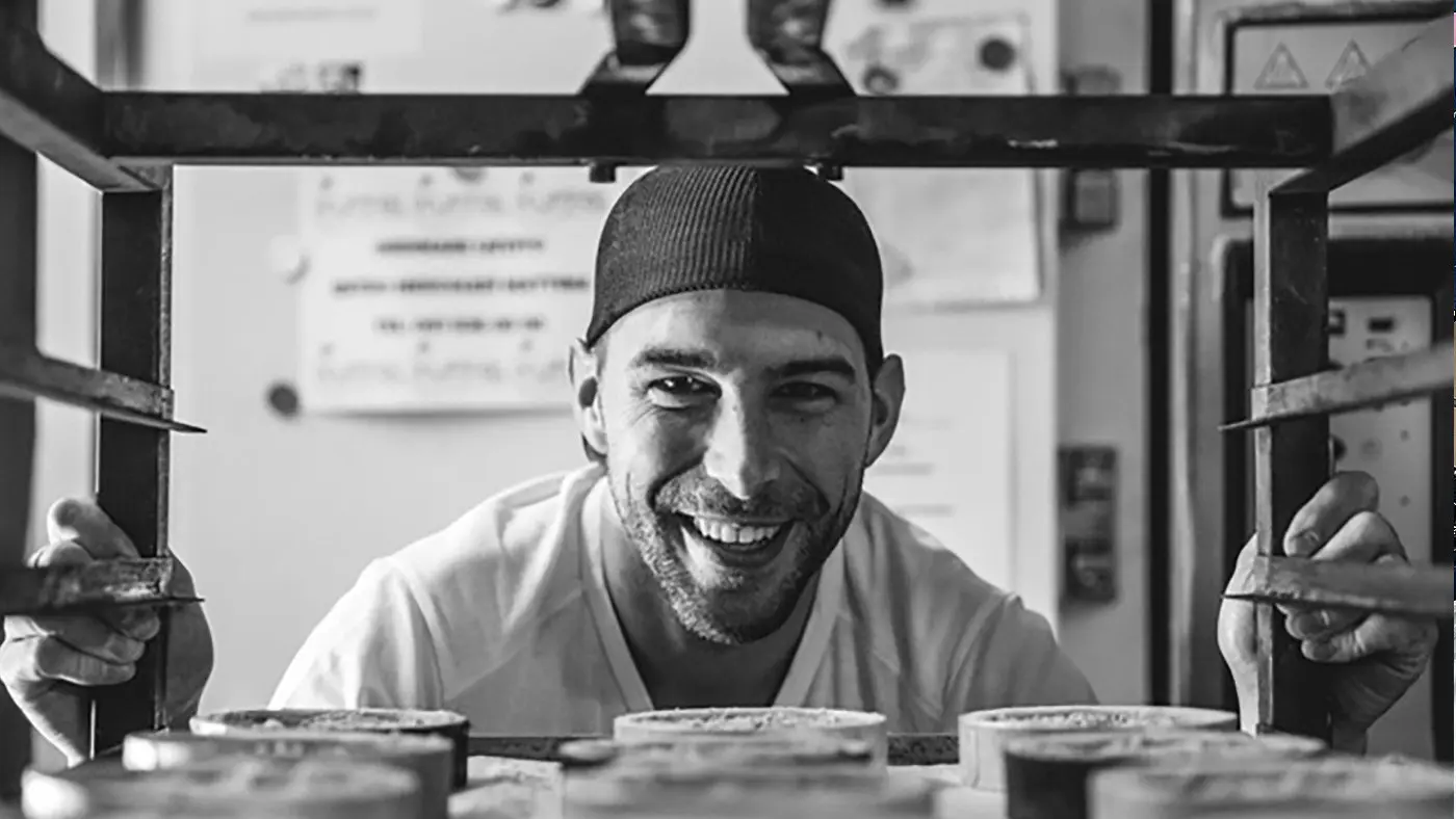 Un uomo sorride mentre guarda attraverso una griglia di un forno. Indossa un berretto e un grembiule, circondato da contenitori di cibo.