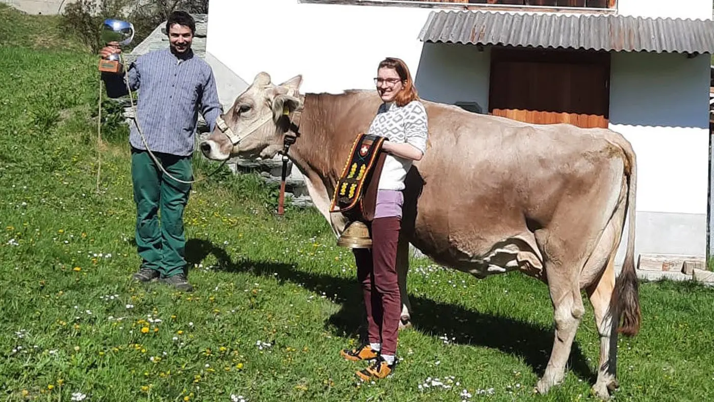 Un uomo tiene un trofeo mentre una donna, sorridente, posa accanto a una mucca in un prato verde. Entrambi indossano abiti casual e sembrano soddisfatti. Sullo sfondo si vede una casa di campagna.