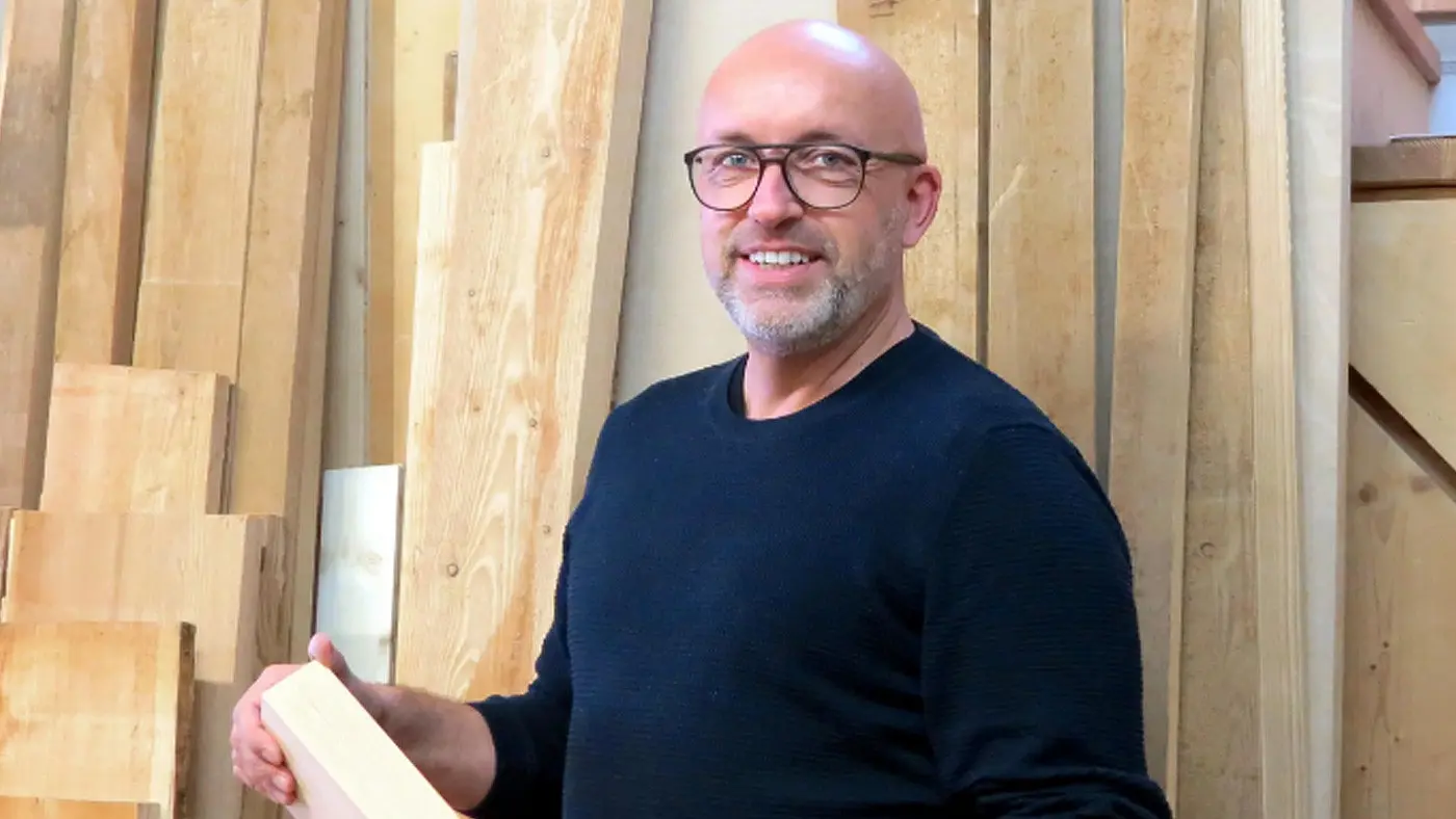 Un uomo calvo con occhiali sorride mentre tiene un pezzo di legno, circondato da pannelli di legno in un ambiente di lavoro.