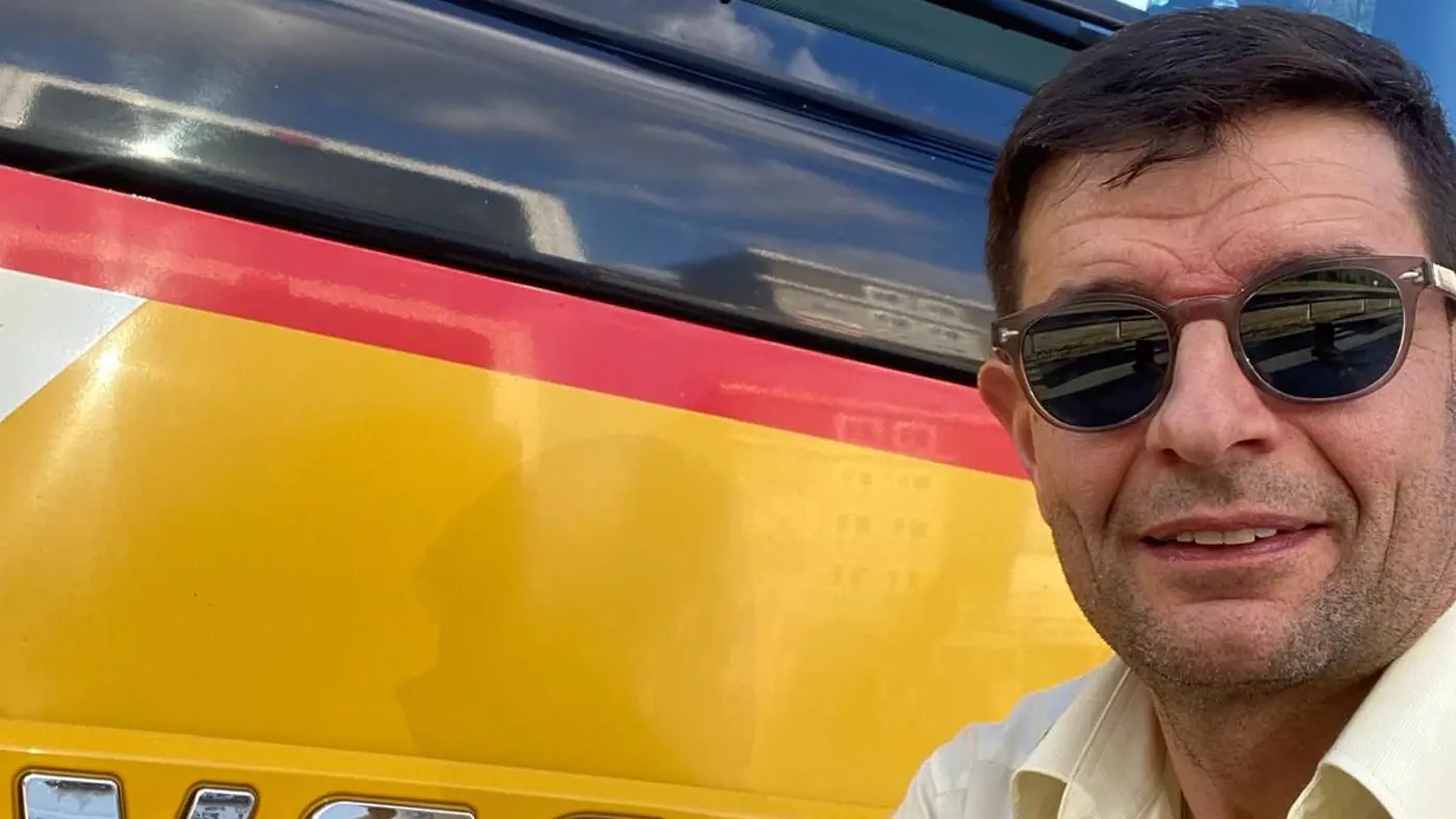 Uomo sorridente con occhiali da sole accanto a un veicolo giallo e rosso. Sullo sfondo si vede un edificio e un cielo blu.