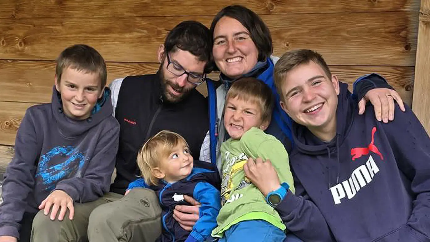 Una famiglia sorridente seduta su una panchina di legno. Ci sono quattro bambini e due adulti. I bambini indossano abiti colorati e si divertono insieme, mentre un adulto abbraccia un bambino più piccolo.