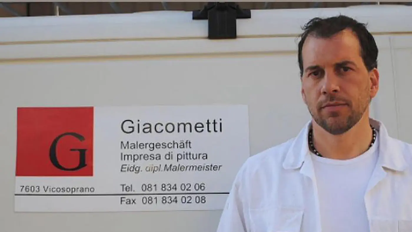 Un uomo in piedi accanto a un cartello di un'azienda di pittura, che mostra il nome "Giacometti" e informazioni di contatto. Indossa una camicia bianca e sembra serio.