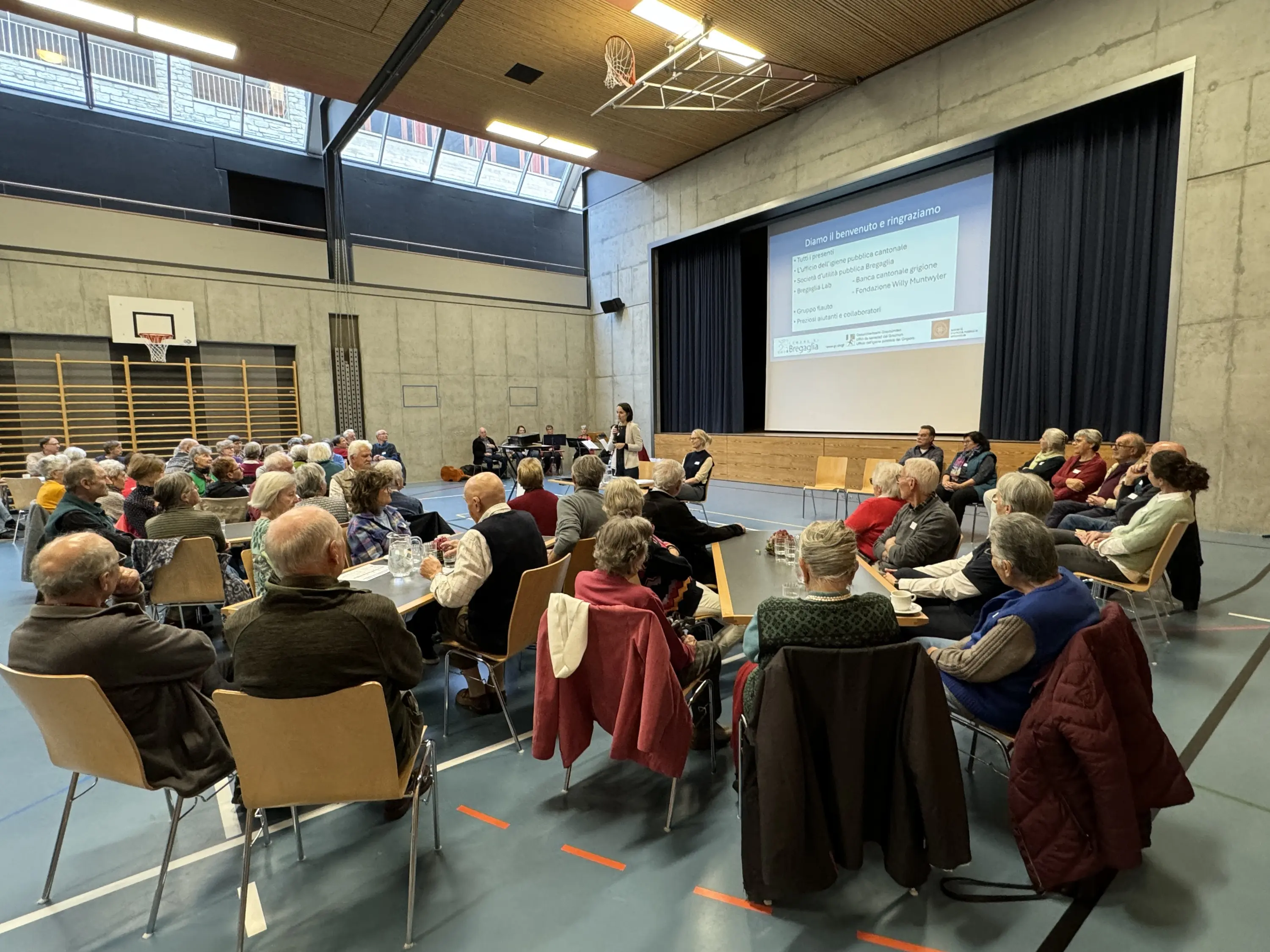 Un gruppo di persone anziane partecipa a un incontro in una palestra. Alcuni relatori sono posizionati su un palco, mentre il pubblico ascolta attentamente. Un proiettore mostra una presentazione sullo schermo.