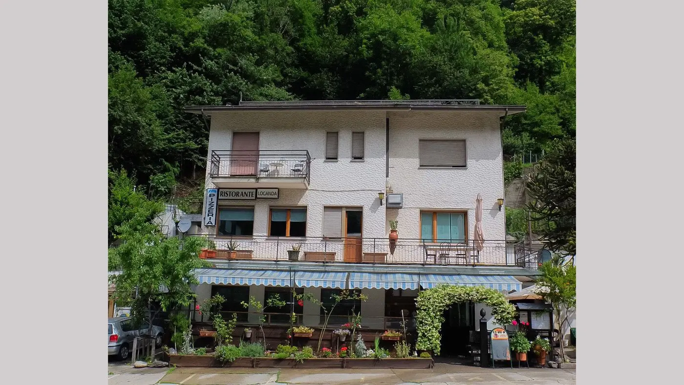Un ristorante a due piani con veranda all'aperto, circondato da alberi verdi. La facciata è bianca con finestre e balconi. È presente una scritta "Ristorante" all'esterno.