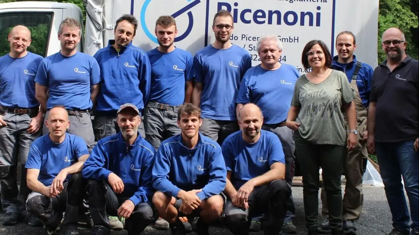 Gruppo di undici persone in maglietta blu, sorridenti, in posa davanti a un grande striscione con il logo "Picenoni". Il gruppo comprende uomini e donne di varie età, tutti vestiti in abbigliamento da lavoro.