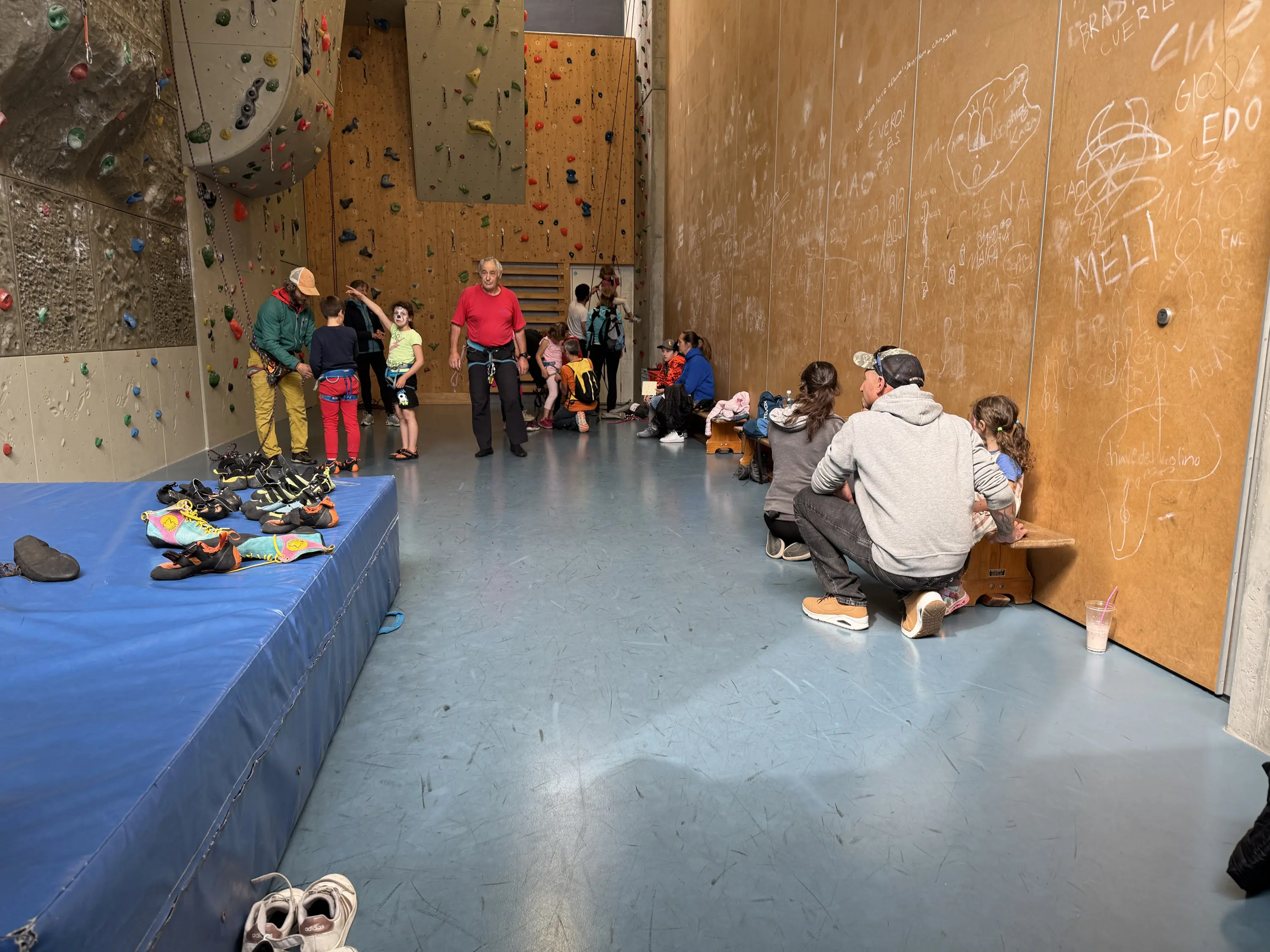 Un muro di arrampicata con diverse persone, tra cui bambini e adulti, che si preparano e si allenano. Alcuni scarpini da arrampicata sono sparsi sul pavimento. Sul fondo, ci sono due persone che assistono bambini mentre indossano l'imbracatura. Le pareti sono decorate con tratti di gesso.
