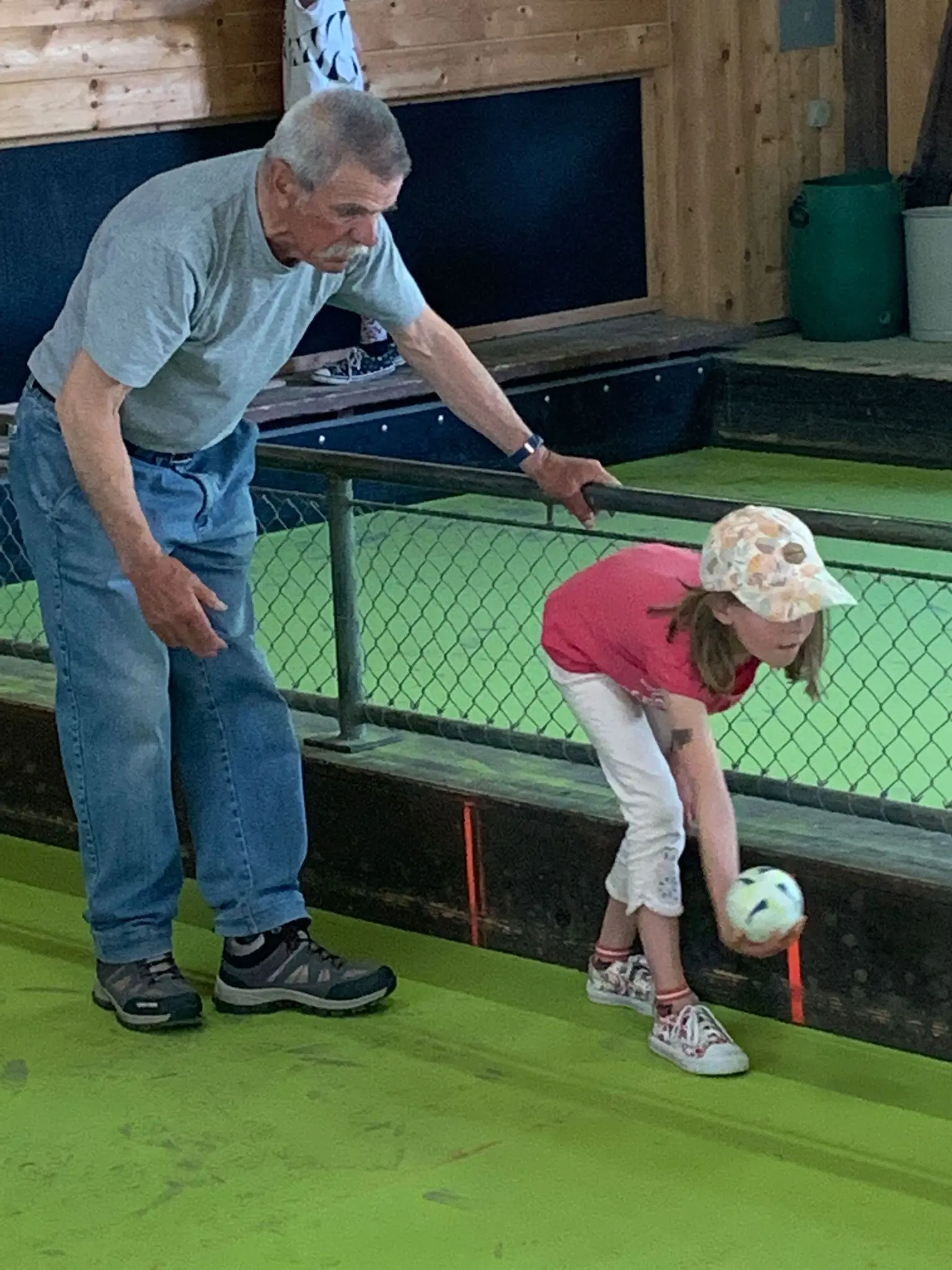 Un uomo anziano aiuta una bambina a giocare a bocce in una pista verde. La bambina indossa una maglietta rosa e un cappello, mentre l'uomo la guida con attenzione.