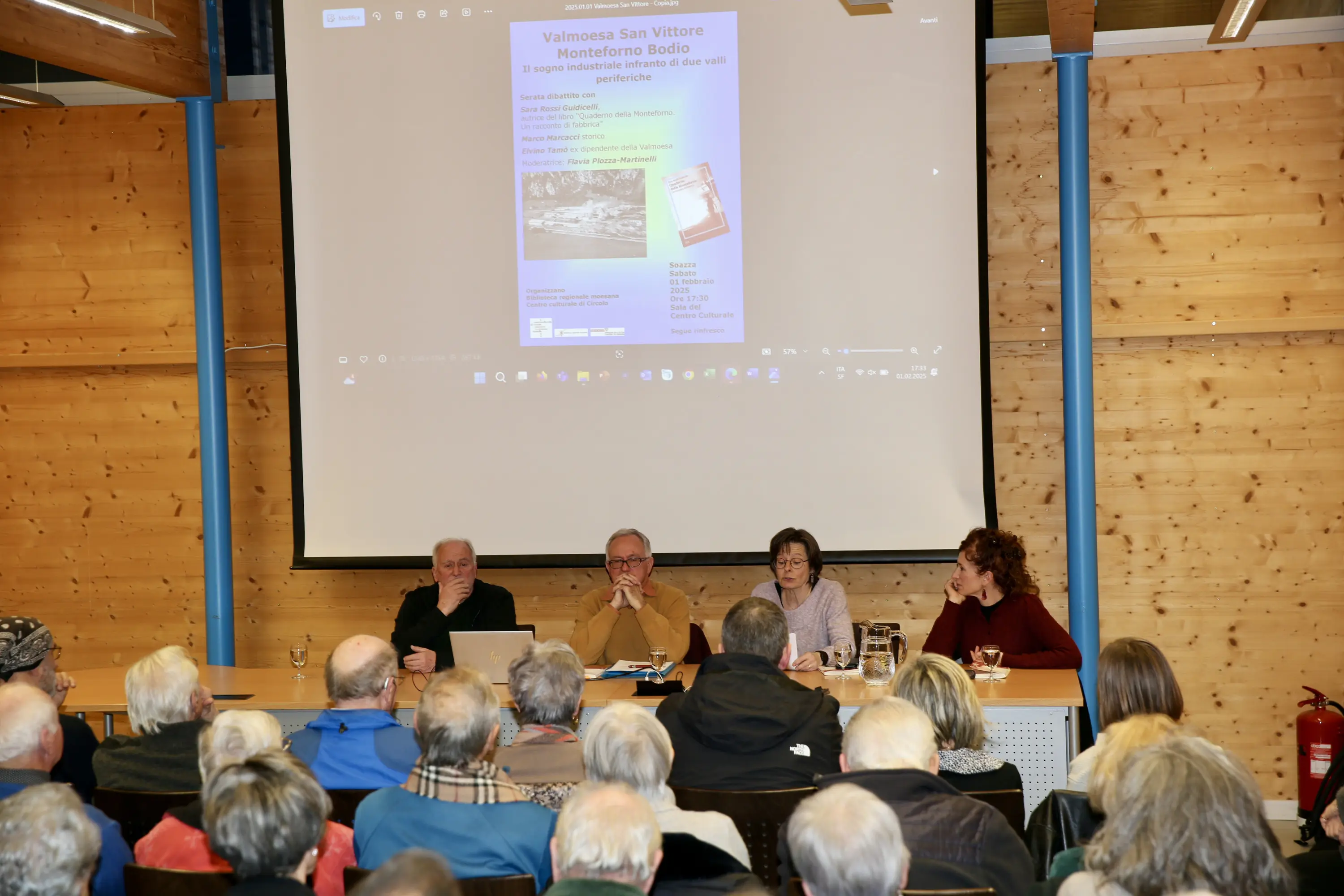Un gruppo di relatori partecipa a un evento pubblico in una sala riunioni. Sullo sfondo è proiettata una presentazione, mentre il pubblico ascolta attentamente.