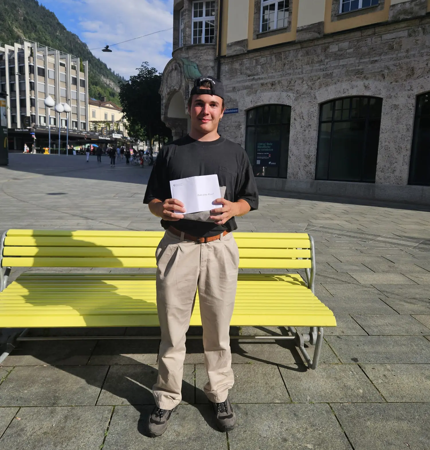 Un giovane uomo in piedi su una panchina gialla, con una maglietta nera e pantaloni chiari. Tiene in mano un foglio bianco e sorride. Sullo sfondo si possono vedere edifici e persone in un'area urbana.