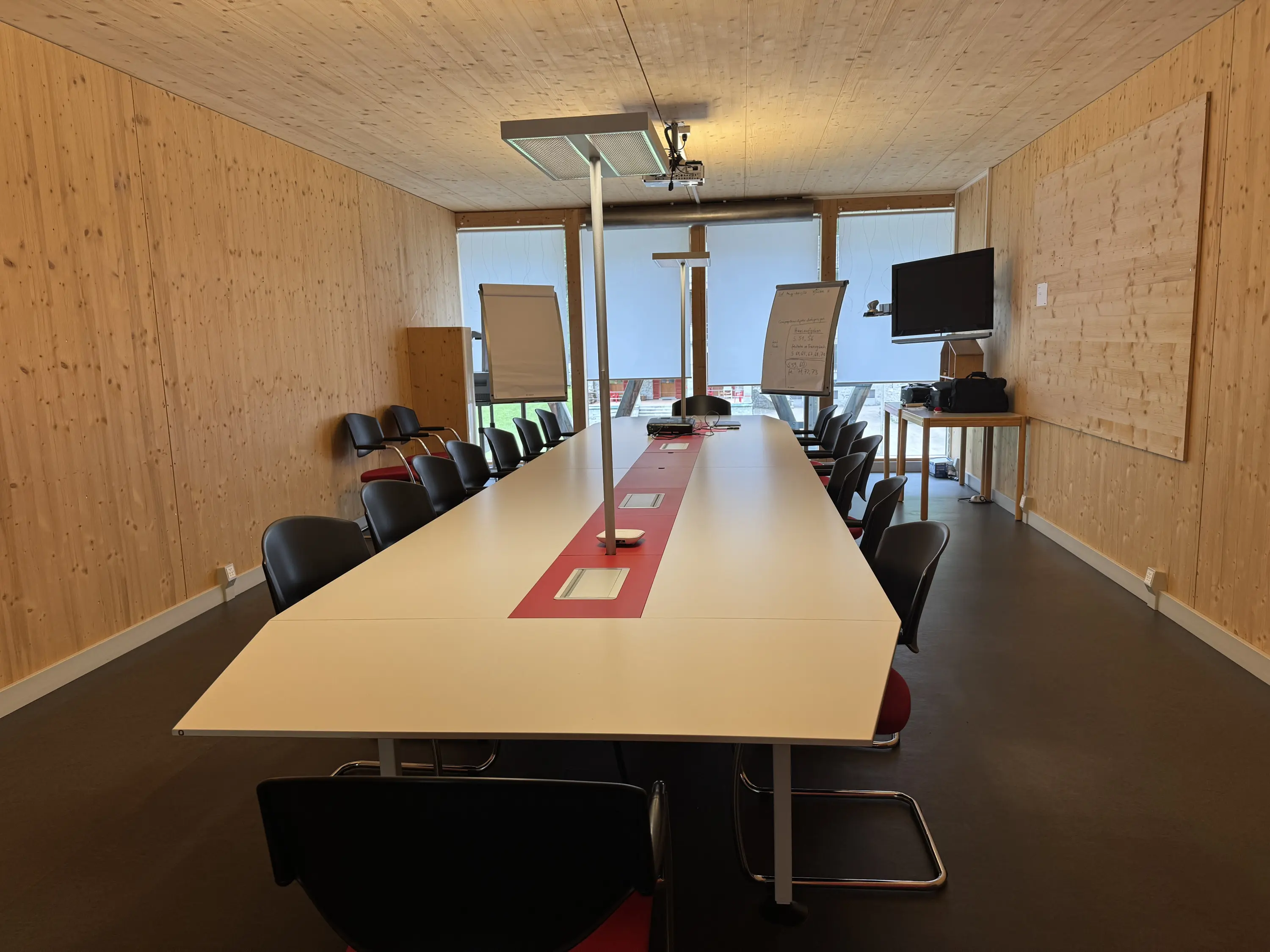 Sala riunioni moderna con una lunga tavola centrale e sedie nere. Pareti in legno chiaro e ampie finestre che offrono una vista esterna. Un proiettore è appeso al soffitto e ci sono strumenti di presentazione visibili.