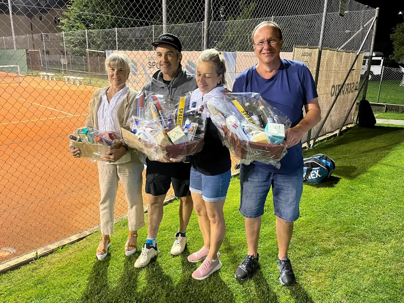 Quattro persone sorridenti tengono in mano cesti di regalo, circondate da una campi da tennis illuminati e una rete.