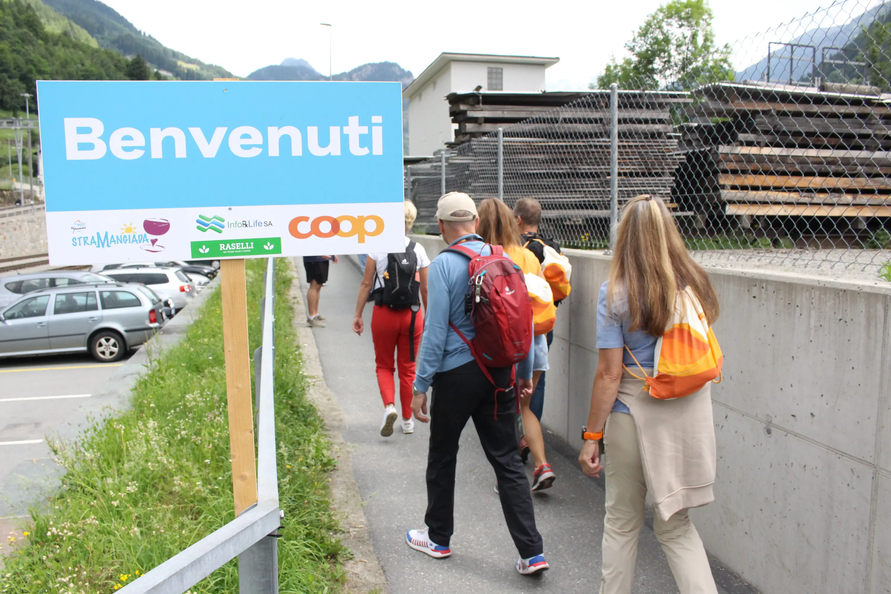 Gruppo di persone che camminano lungo un sentiero, con un cartello che dice "Benvenuti". Sullo sfondo si vedono auto e strutture in legno.