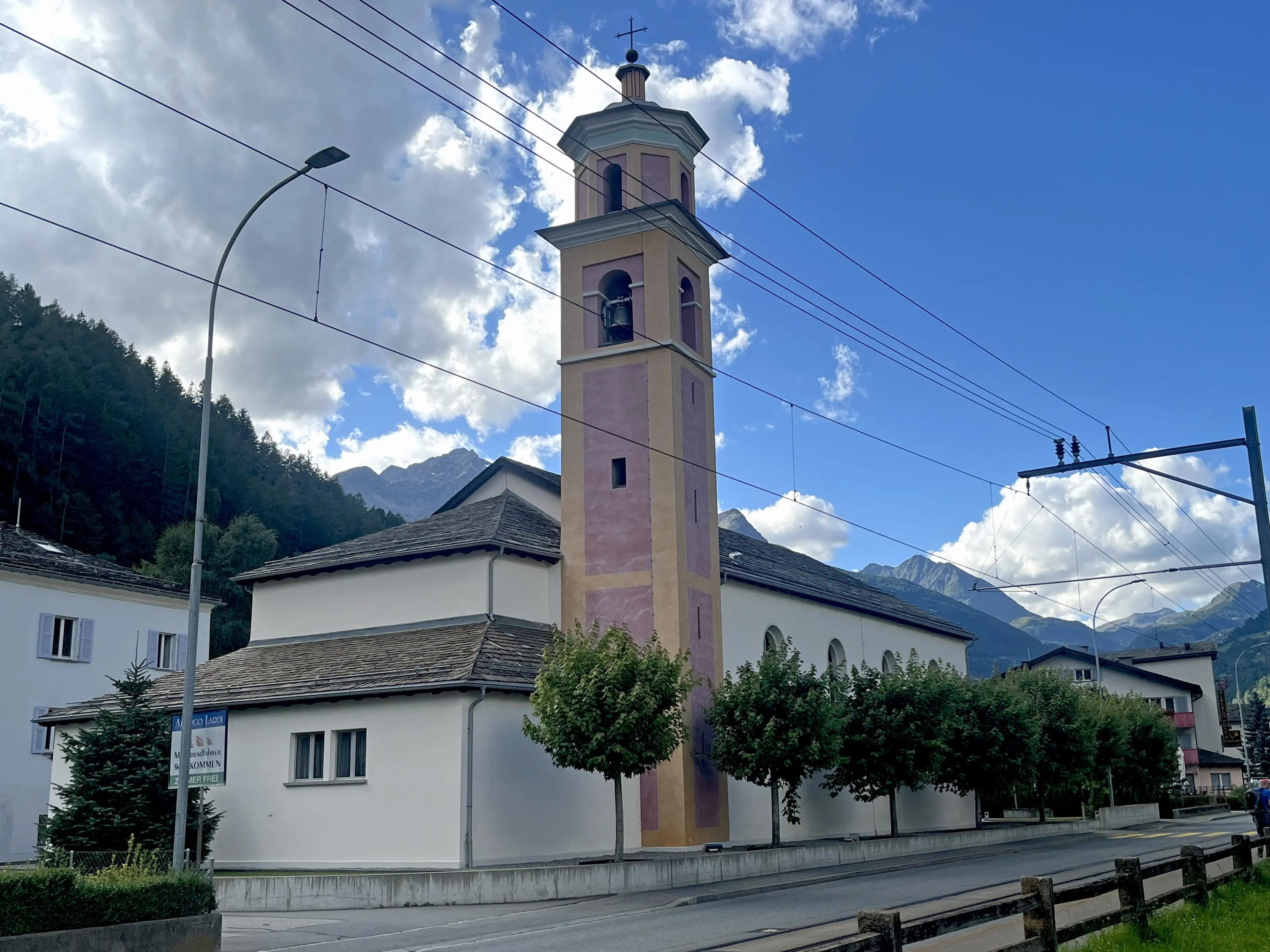 Chiesa di San Francesco d’Assisi a Le Prese in Valposchiavo con torre campanaria rosa e bianca, circondata da alberi e montagne sullo sfondo. Il cielo è parzialmente nuvoloso.