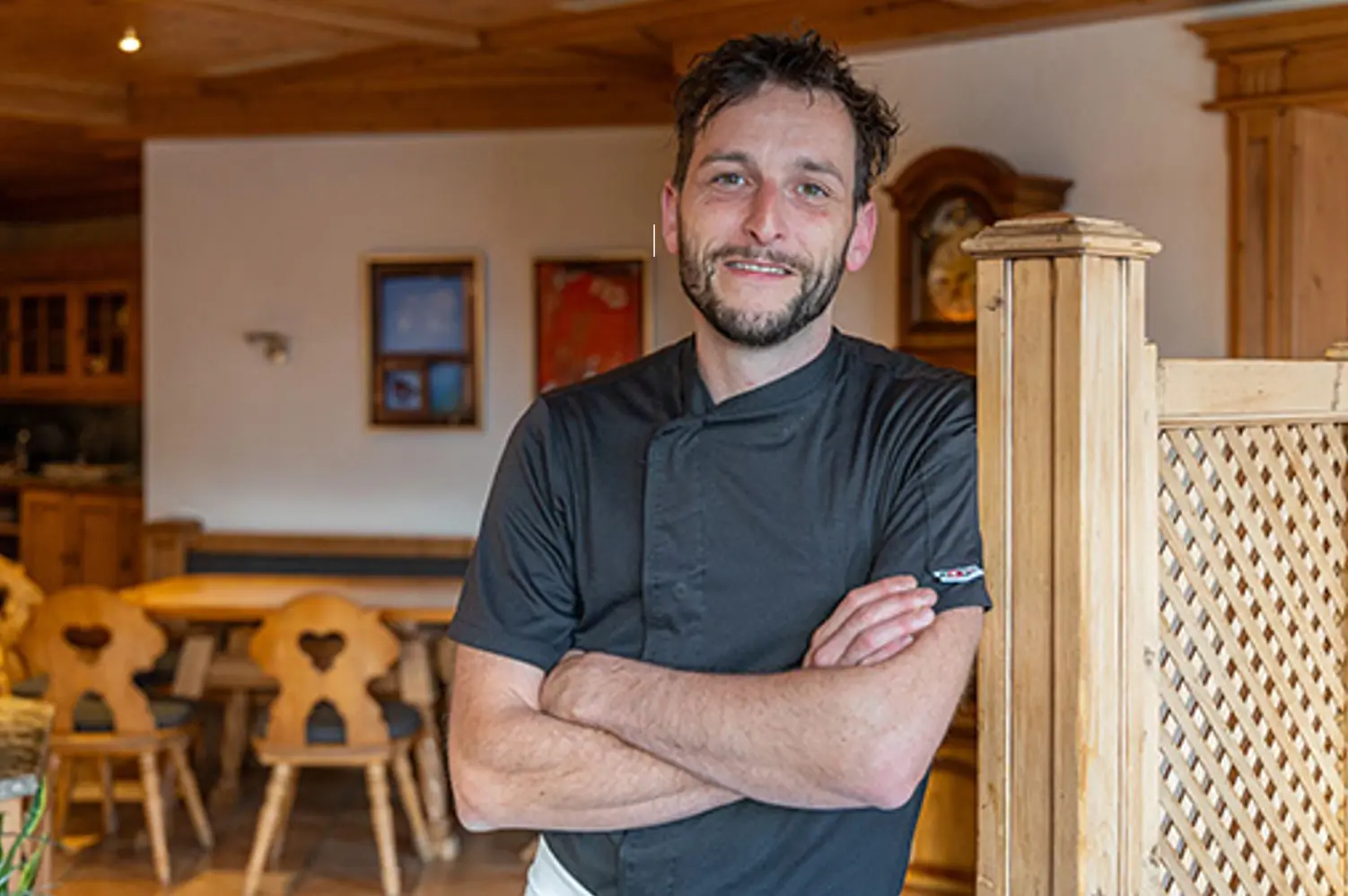 Un cuoco sorridente indossa una giacca da cucina nera, con le braccia incrociate, mentre si trova in un ristorante accogliente con arredi in legno. Si intravedono tavoli e sedie tipiche sullo sfondo.