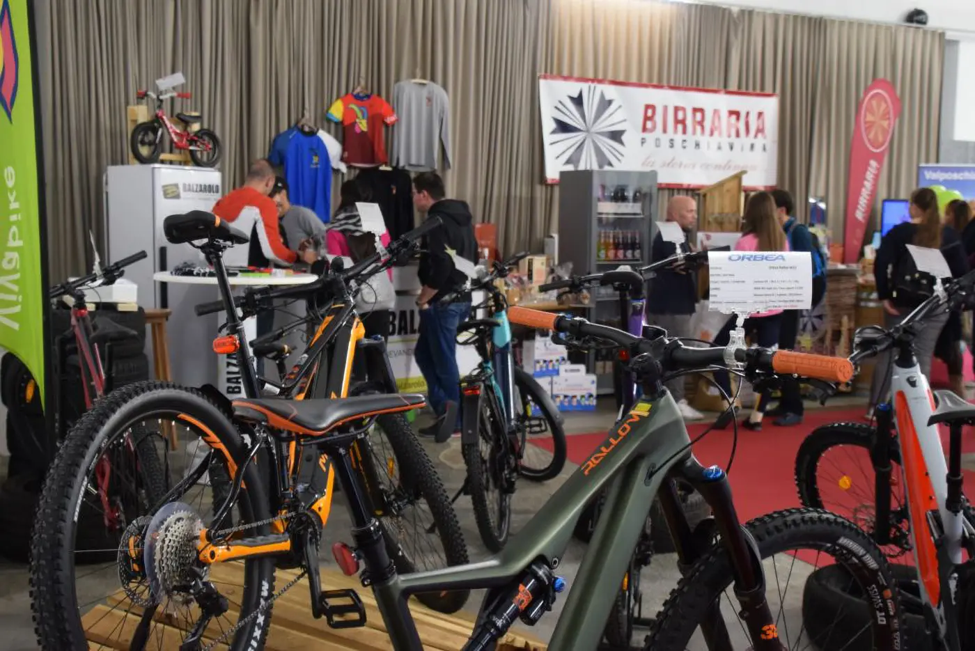 Un'esposizione di biciclette con vari modelli in primo piano, tra cui mountain bike. Si vedono visitatori e stand con prodotti e abbigliamento sportivo sullo sfondo.