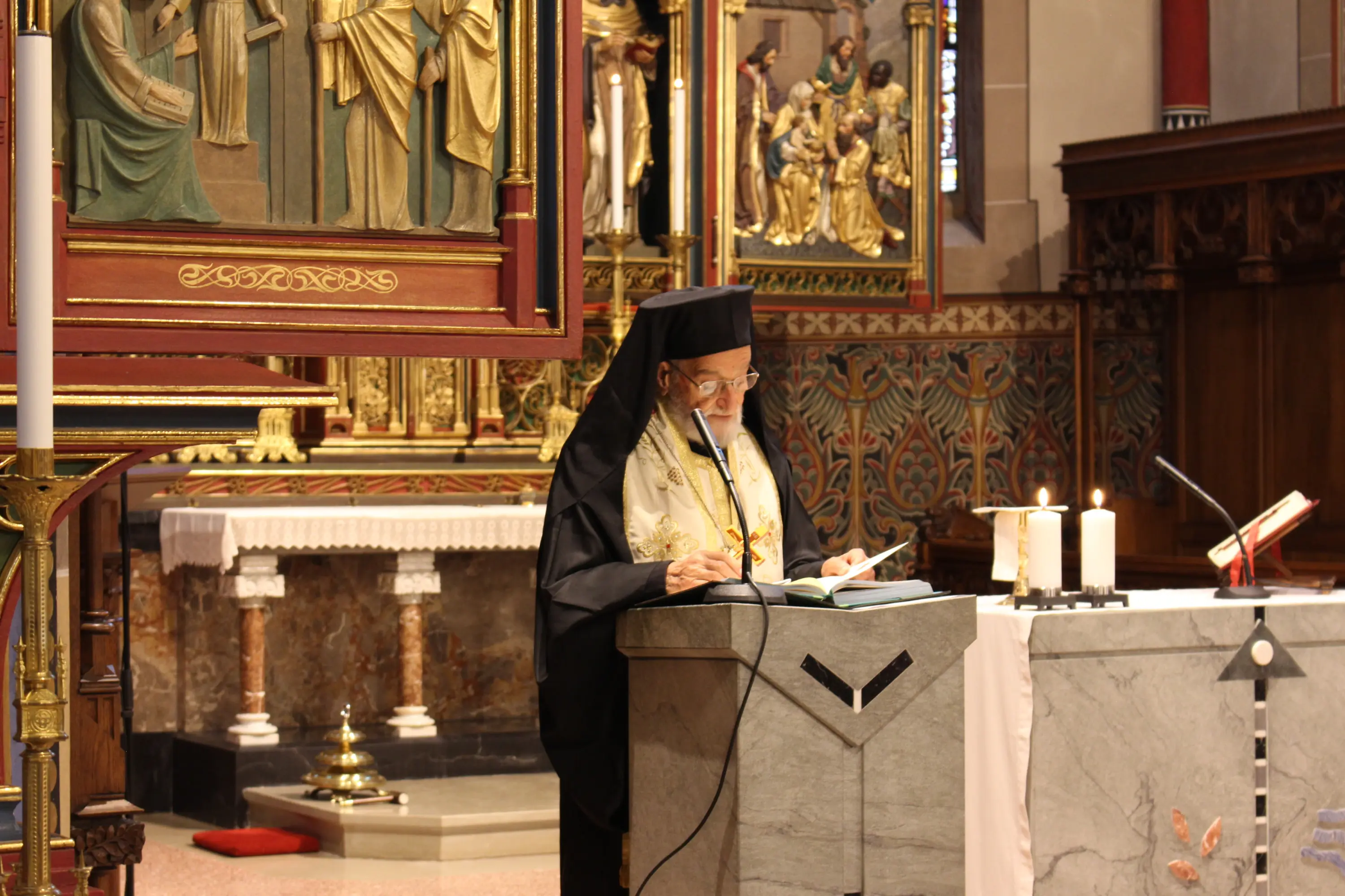 Un uomo in abiti religiosi, con un copricapo scuro, legge da un libro dietro un altare in una chiesa decorata, con statue e candele in evidenza.