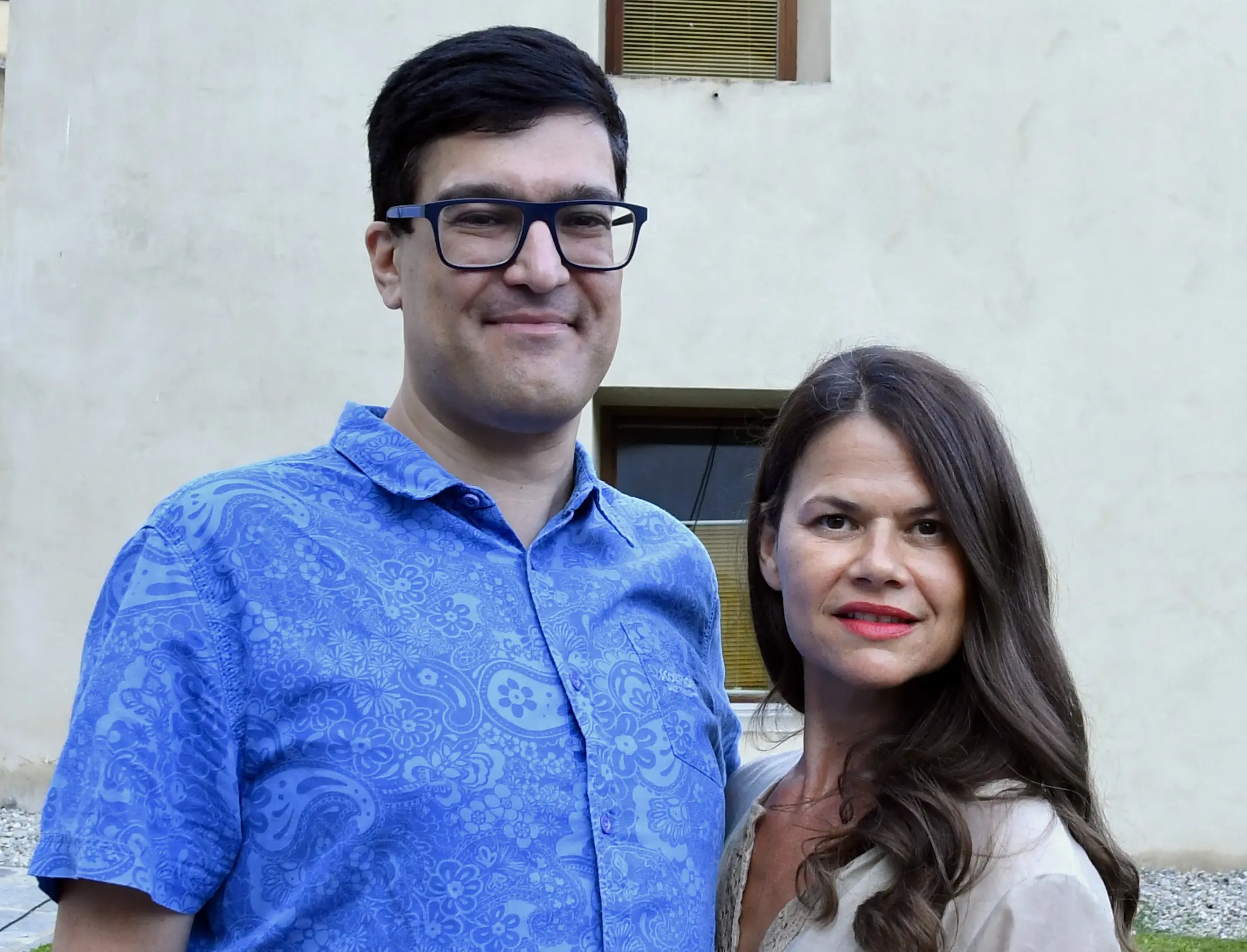 Una coppia sorridente in piedi davanti a una parete chiara. L'uomo indossa una camicia a fantasia blu, mentre la donna ha i capelli lunghi e indossa una blusa chiara.