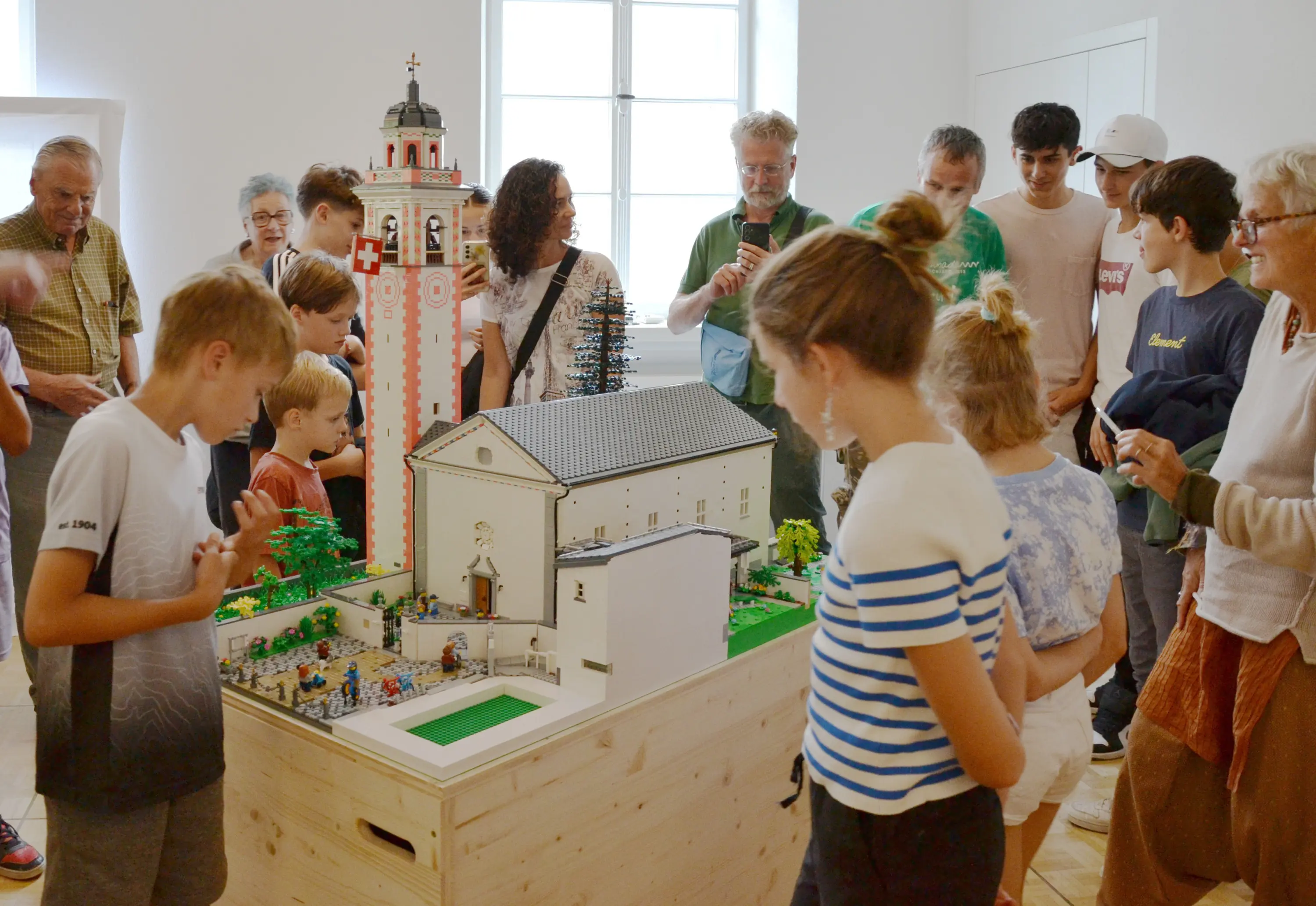 Un gruppo di persone di diverse età osserva una diorama di Lego che rappresenta un edificio storico, circondato da dettagli in miniatura e alberi, all'interno di un museo.