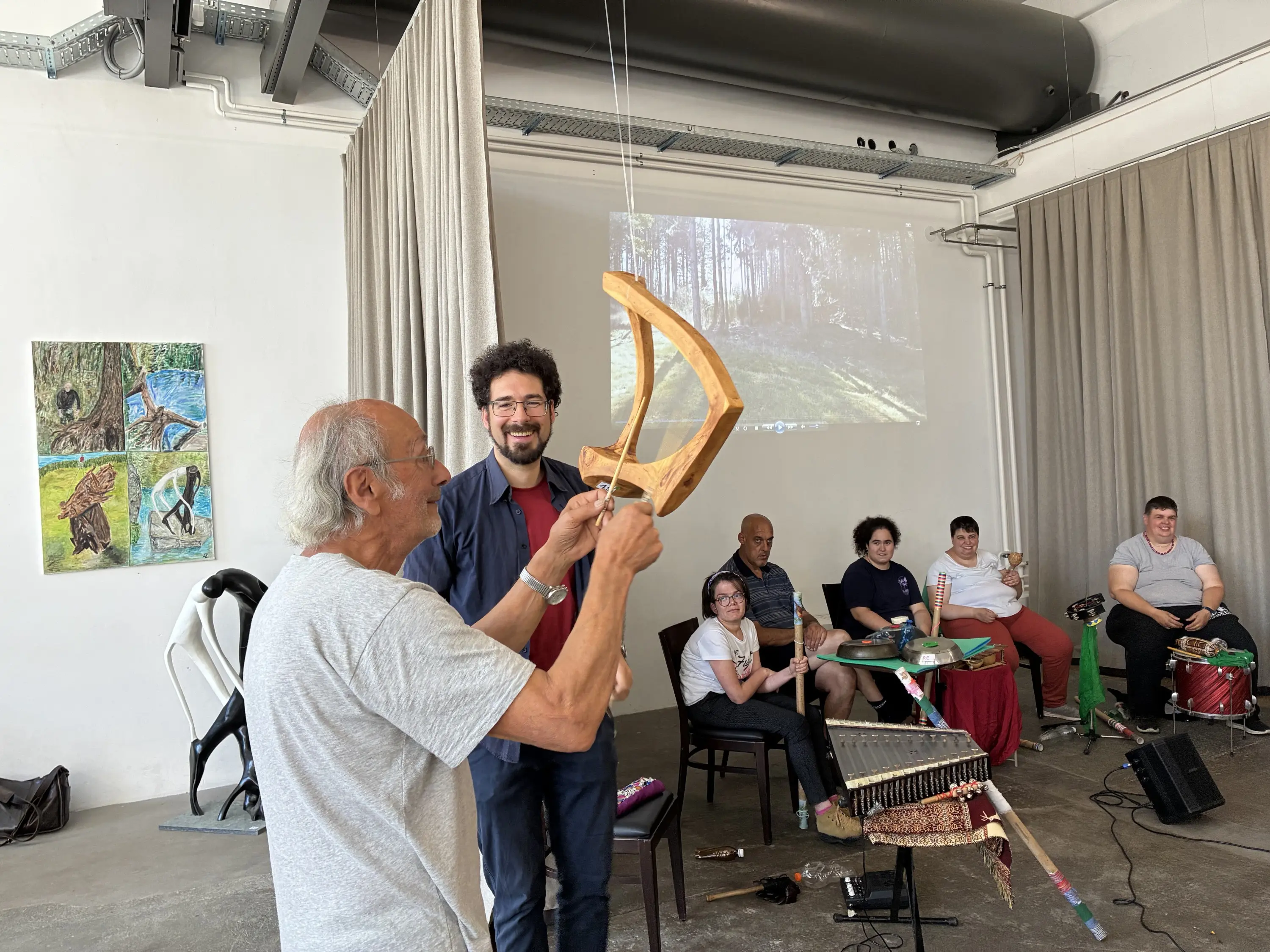 Un uomo anziano mostra un arco musicale a un giovane in un ambiente di laboratorio, mentre un gruppo di persone assiste. Sullo sfondo, un'immagine proiettata di un bosco.