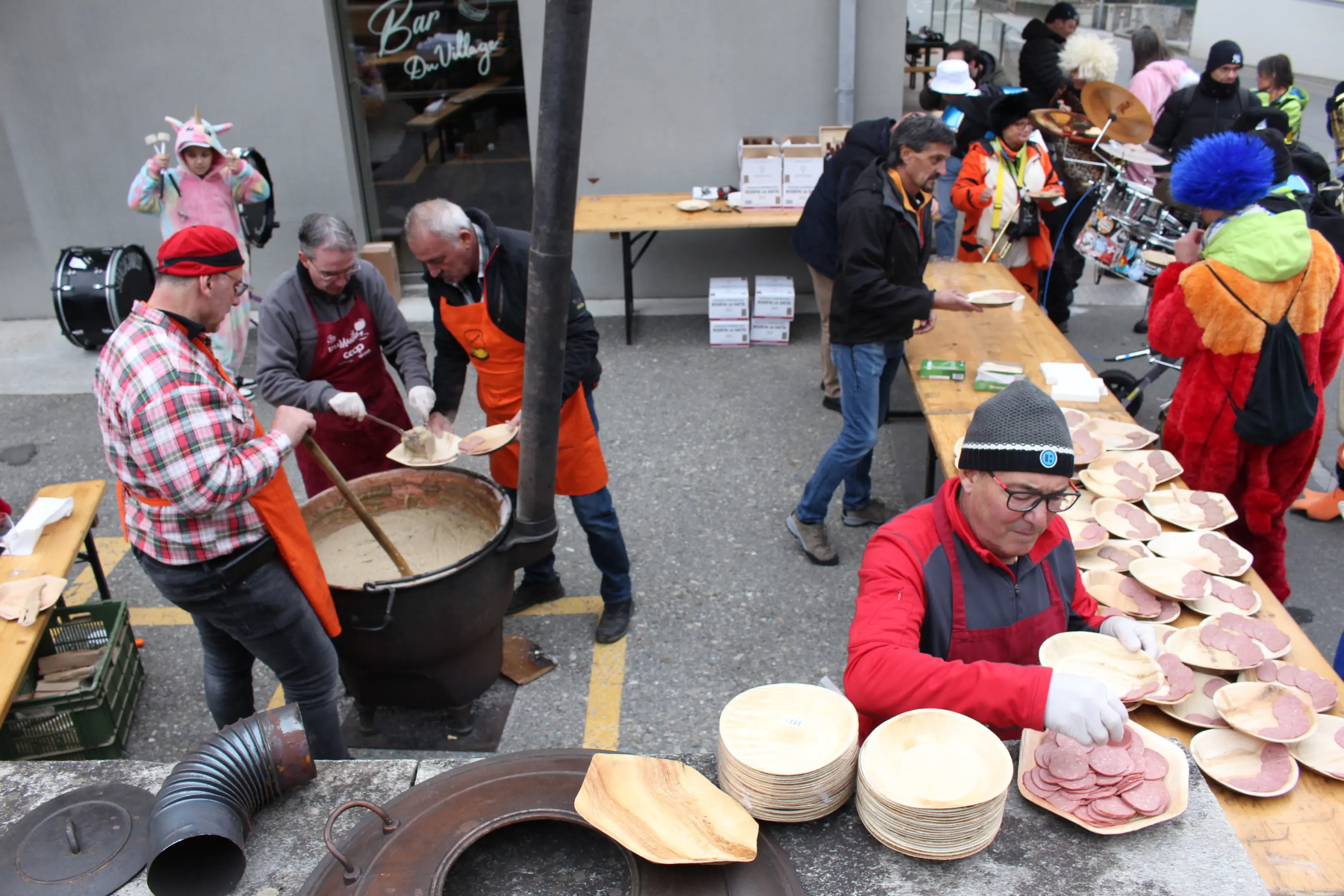 Un gruppo di persone sta preparando piatti di legno con cibo, mentre alcuni indossano grembiuli arancioni. Un grande pentolone è sul fuoco, e si vedono anche membri di una banda musicale sullo sfondo.