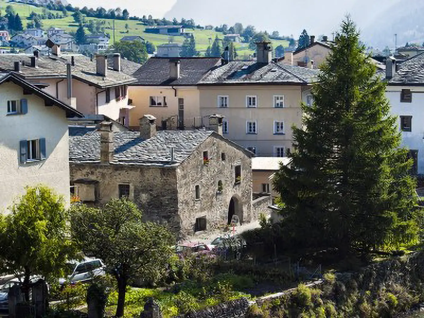 Panorama di un villaggio montano con edifici storici e tetti in pietra. A destra, si erge un albero di conifera. Sullo sfondo, verdi colline e una leggera foschia.