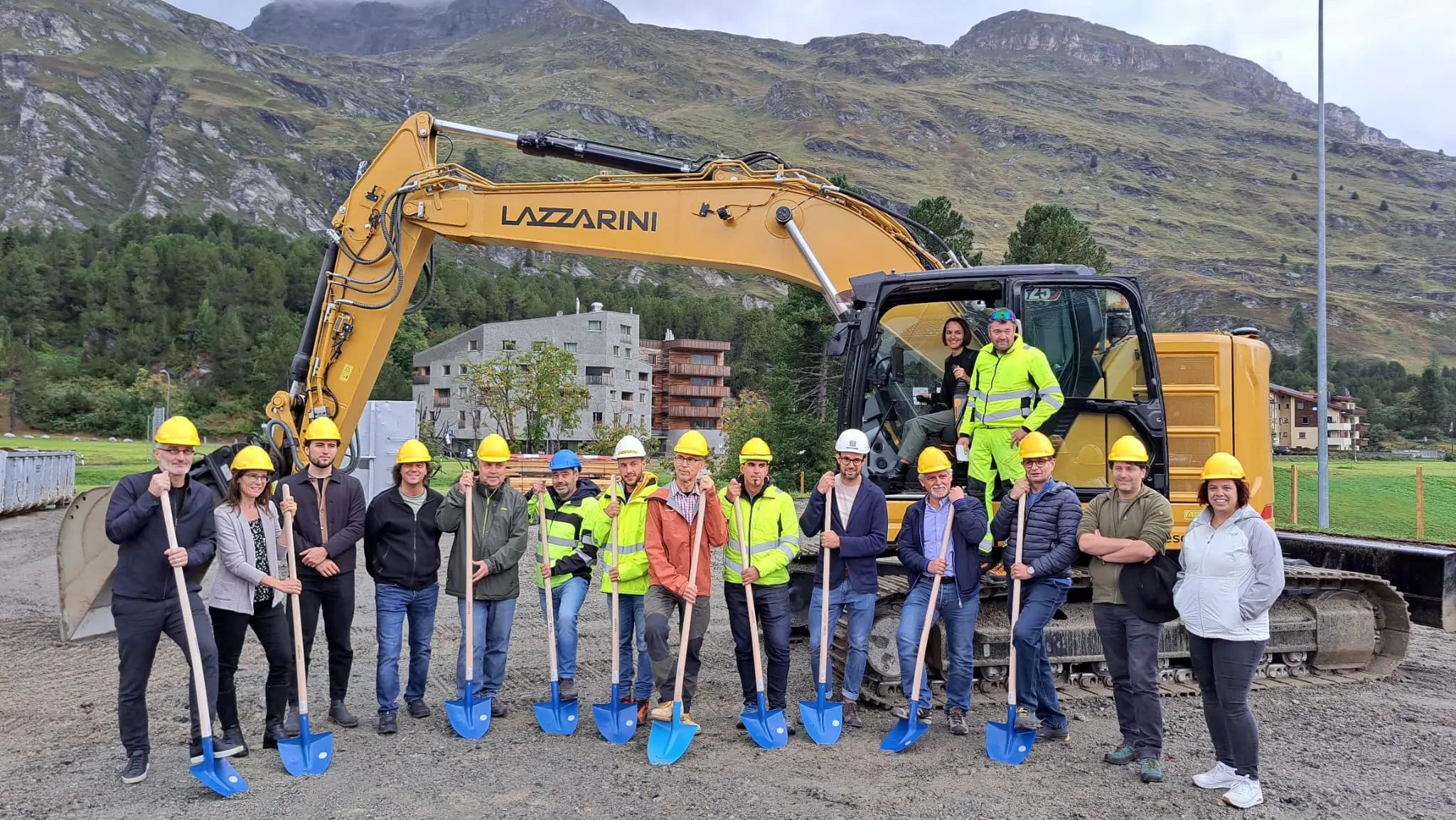 Un gruppo di persone, tutte con elmetti gialli, posa con pala alla mano davanti a un escavatore giallo. Sullo sfondo, si vedono edifici e una montagna verde. È una cerimonia di inaugurazione.