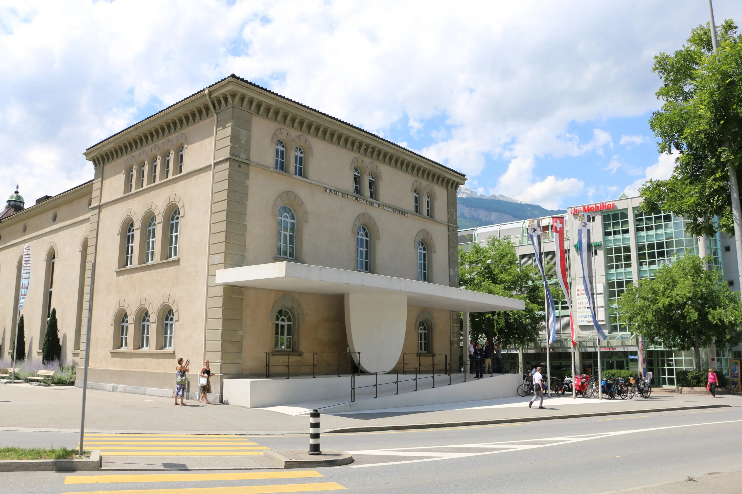Una facciata di un edificio storico con architettura classica, dotata di un ampio portico sostenuto da una struttura moderna. Si trova accanto a un moderno edificio di vetro, circondato da alberi e strade. Persone passeggiano vicino al marciapiede.