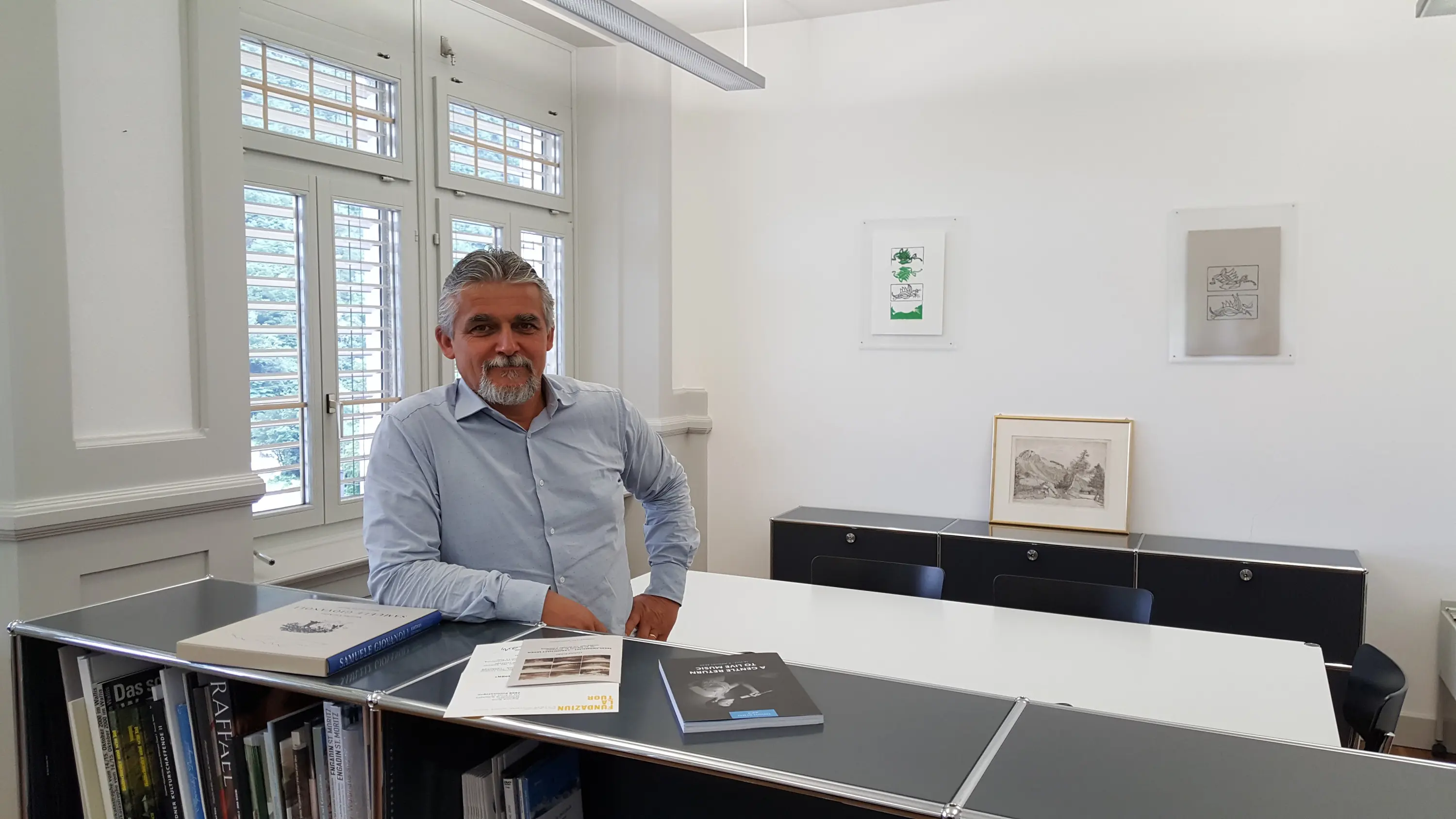 Uomo sorridente con camicia azzurra si appoggia a un tavolo in un ufficio luminoso. Sul tavolo ci sono alcuni libri e documenti. Sullo sfondo, finestre con persiane e opere d'arte appese alle pareti.