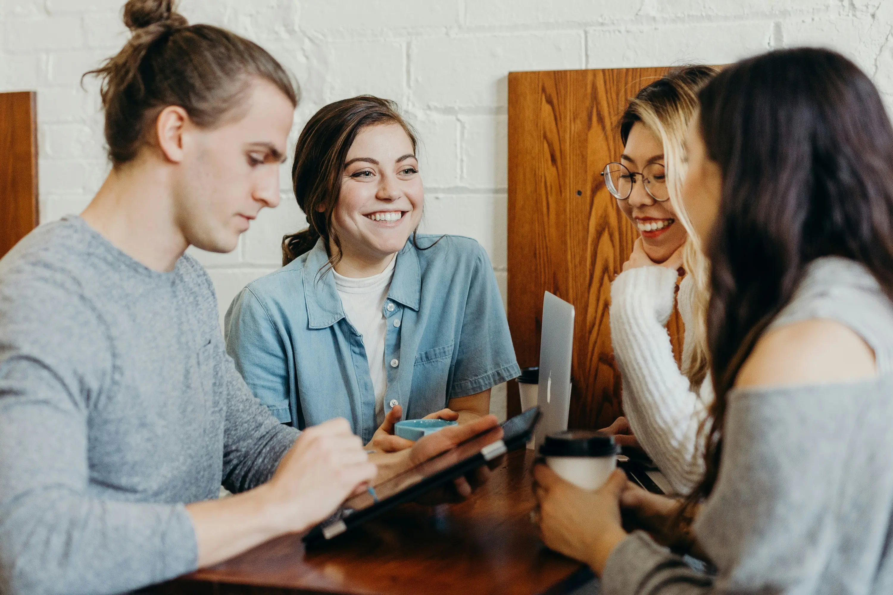 Un gruppo di quattro giovani seduti intorno a un tavolo, sorridendo e conversando. Uno di loro sta usando un tablet, mentre gli altri tengono in mano tazze di caffè. L'atmosfera è informale e amichevole.