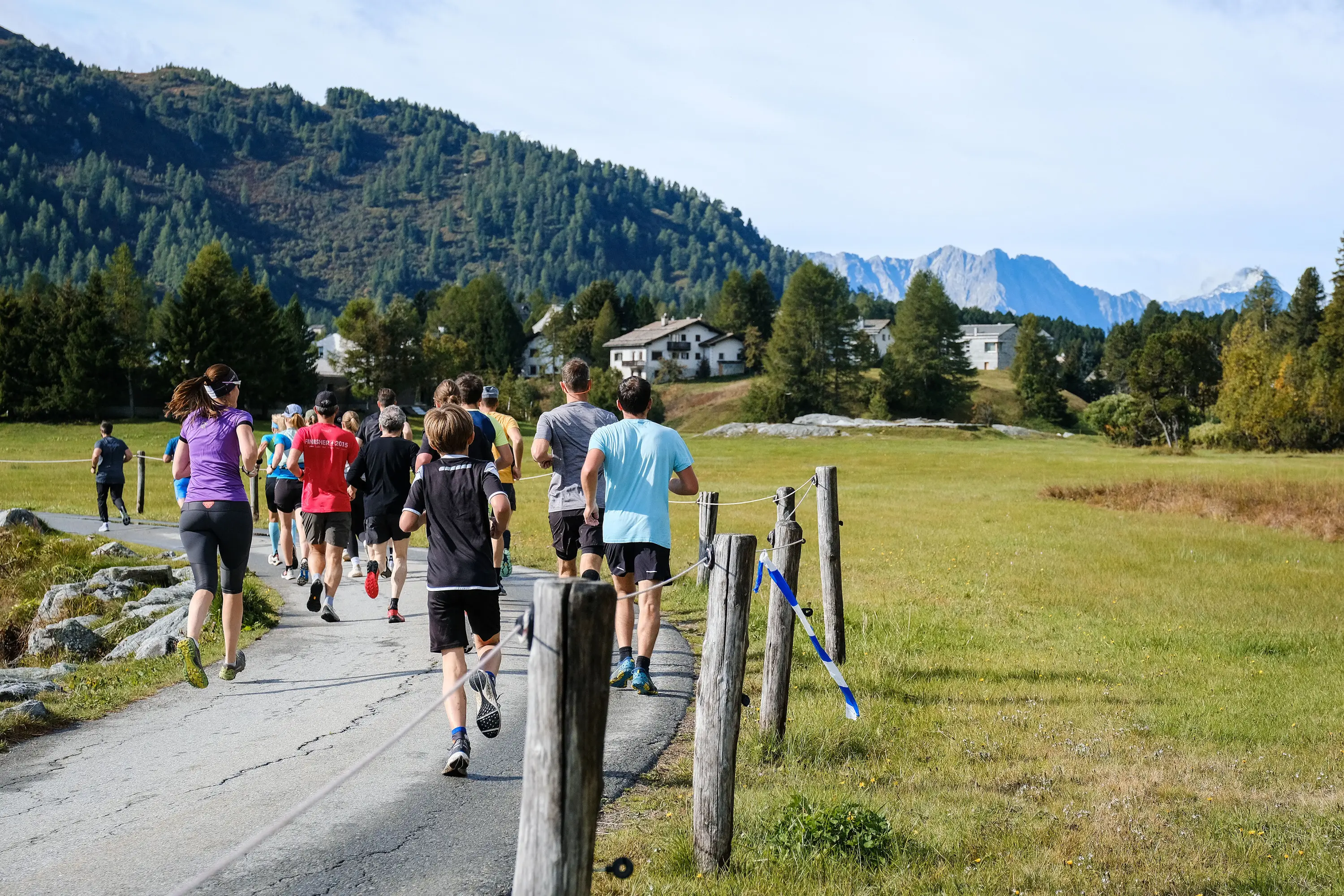 Un gruppo di corridori percorre un sentiero circondato da alberi e montagne. È una giornata soleggiata e il paesaggio è verde e collinare.