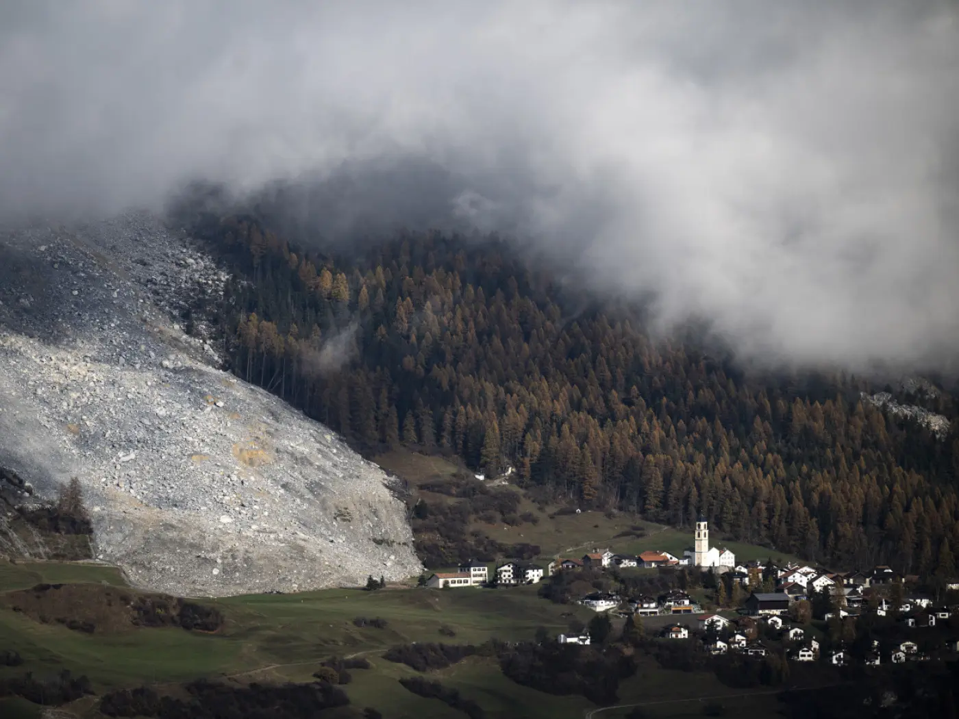 Un paesaggio montano con una frana visibile sul lato sinistro. Nella parte inferiore, un villaggio con case bianche e una chiesa con un campanile è circondato da alberi di conifere e nuvole basse.