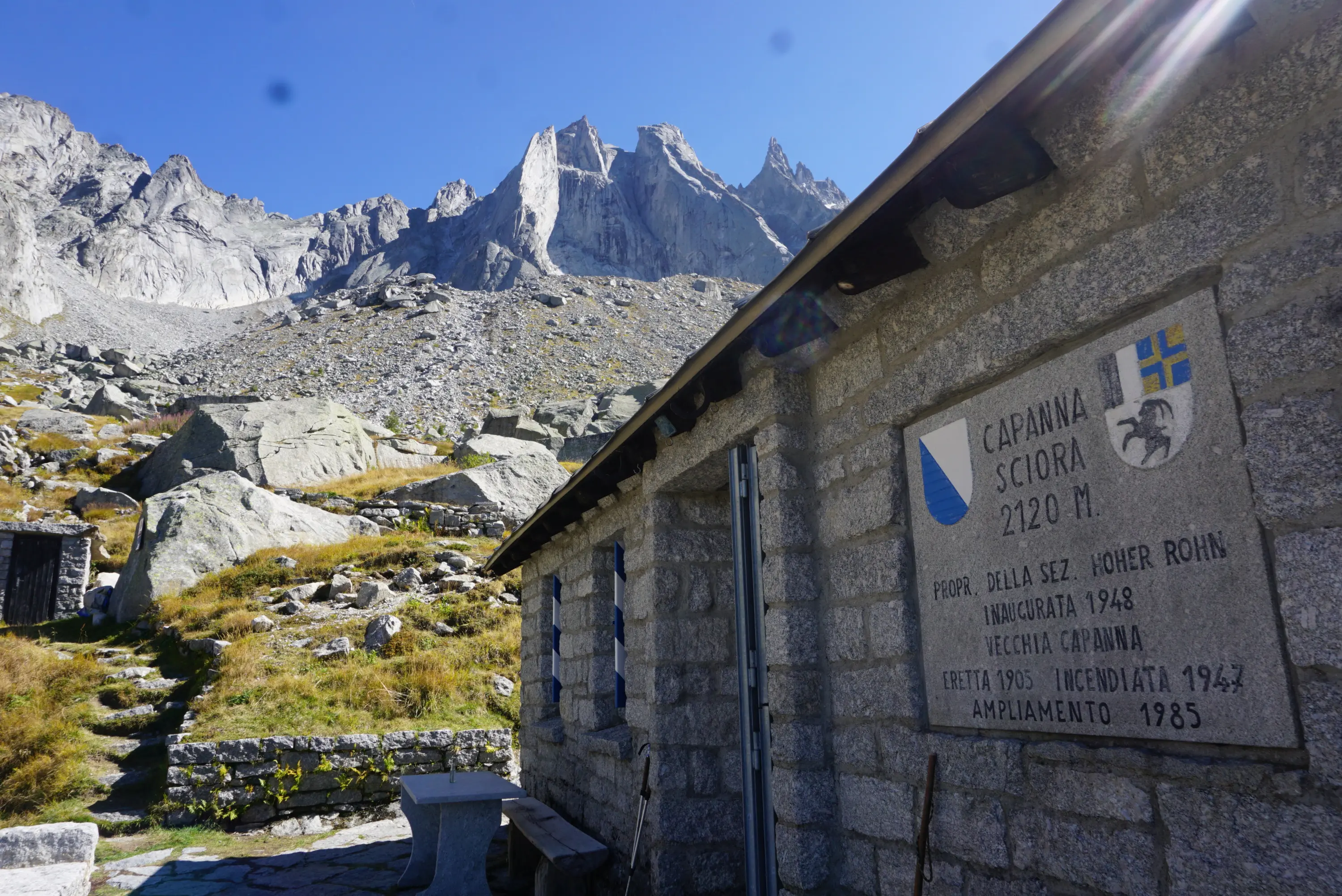 Capanna Sciore a 2120 metri, con un muro in pietra che presenta una targa commemorativa. Sullo sfondo si ergono maestose montagne. La scena è illuminata da un cielo sereno e soleggiato.