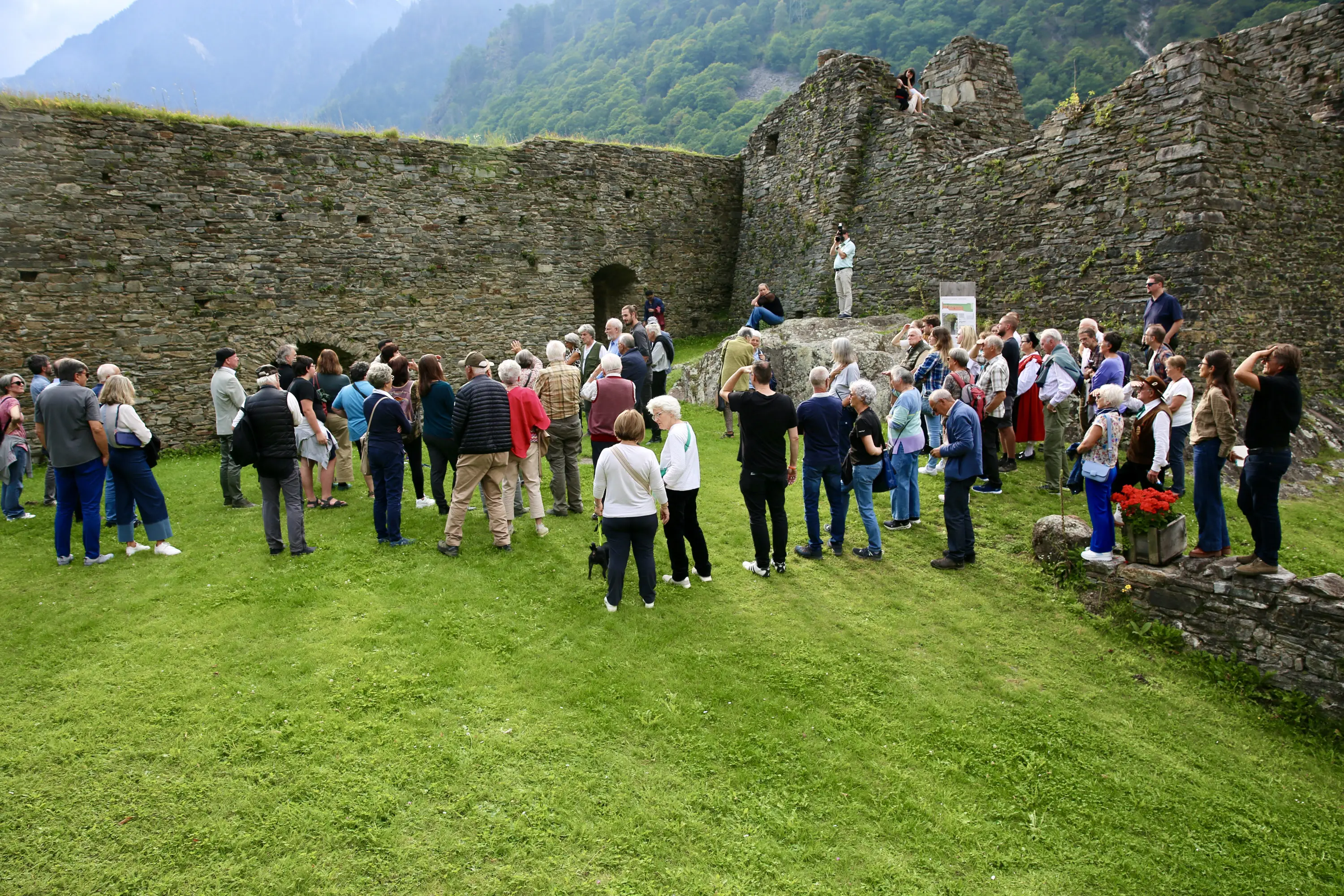 Gruppo di persone radunate in un vasto prato verde davanti a rovine di un antico castello, con montagne sullo sfondo.