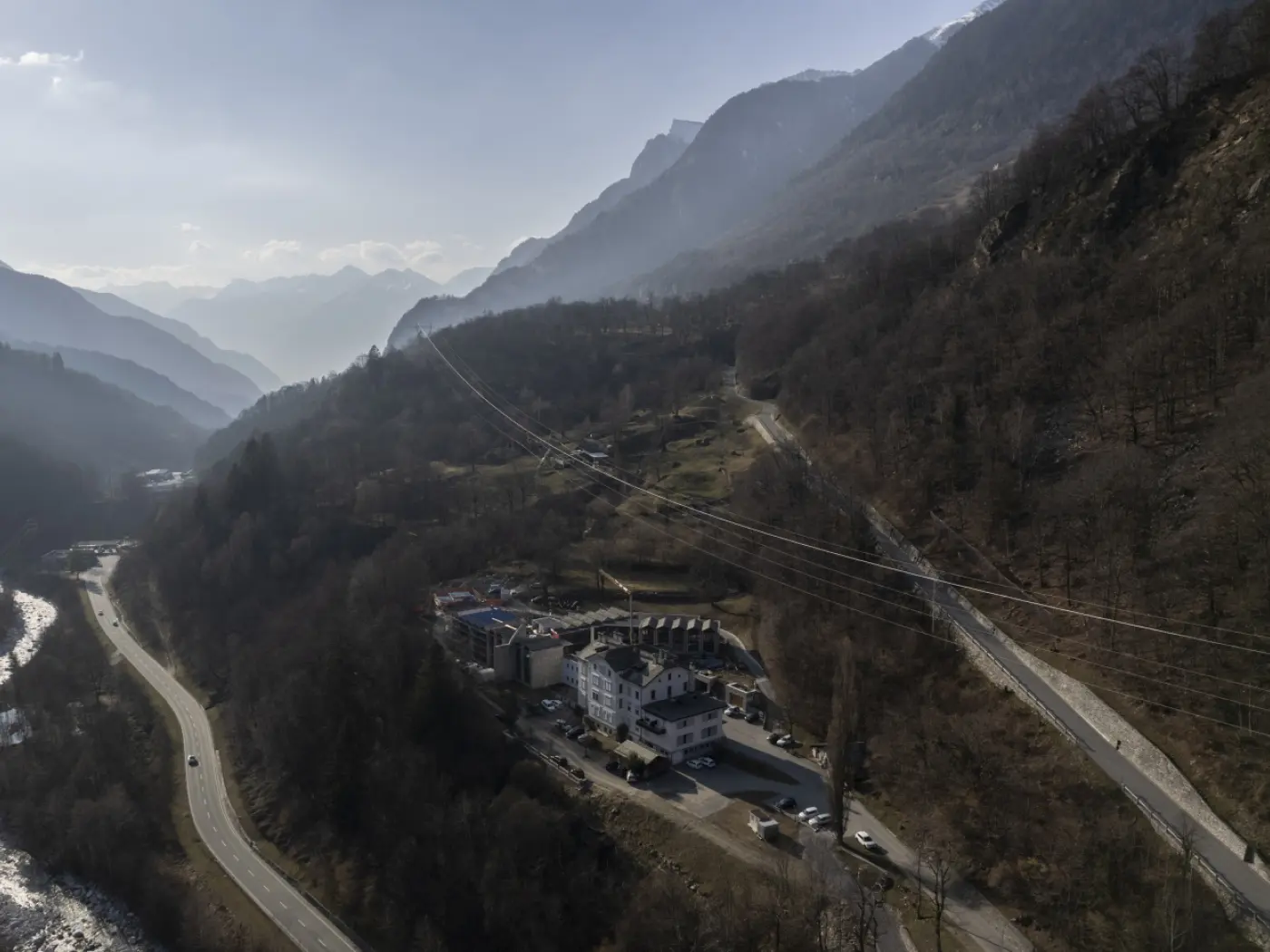 Vista panoramica di un paesaggio montano con una valle, circondata da montagne e un fiume. In primo piano si intravede un complesso alberghiero e strade che si snodano tra gli alberi. La scena è illuminata da una luce soffusa, creando un'atmosfera tranquilla.