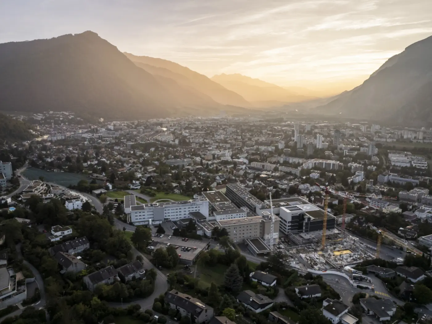 Veduta aerea di una città circondata da monti al tramonto, con edifici residenziali e commerciali visibili, e un cielo sfumato di arancione e blu.