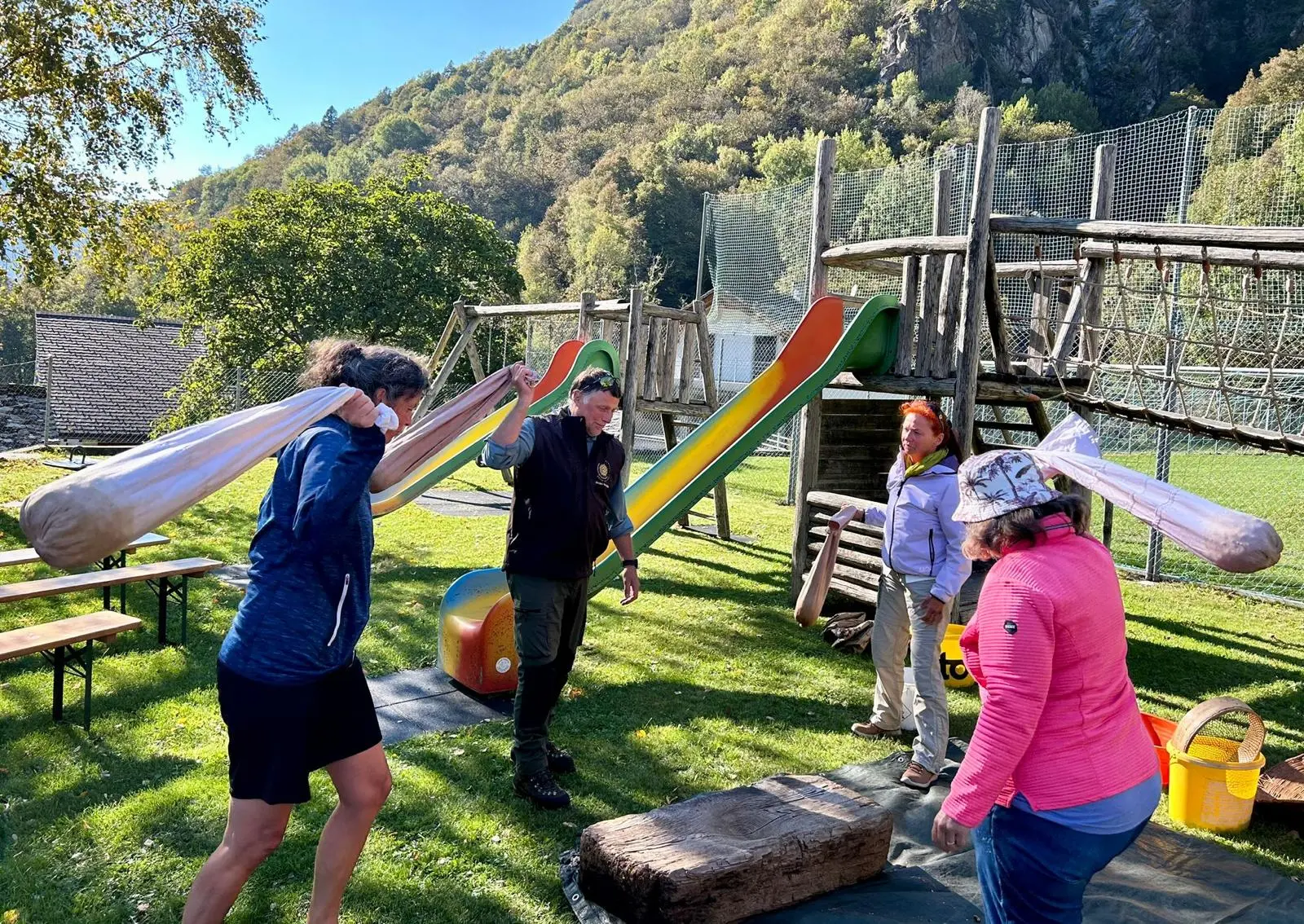 Un gruppo di persone all'aperto partecipa a un'attività fisica, sollevando sacchi. Dietro di loro si trovano scivoli e un'area giochi circondata da alberi e colline. È una giornata di sole.