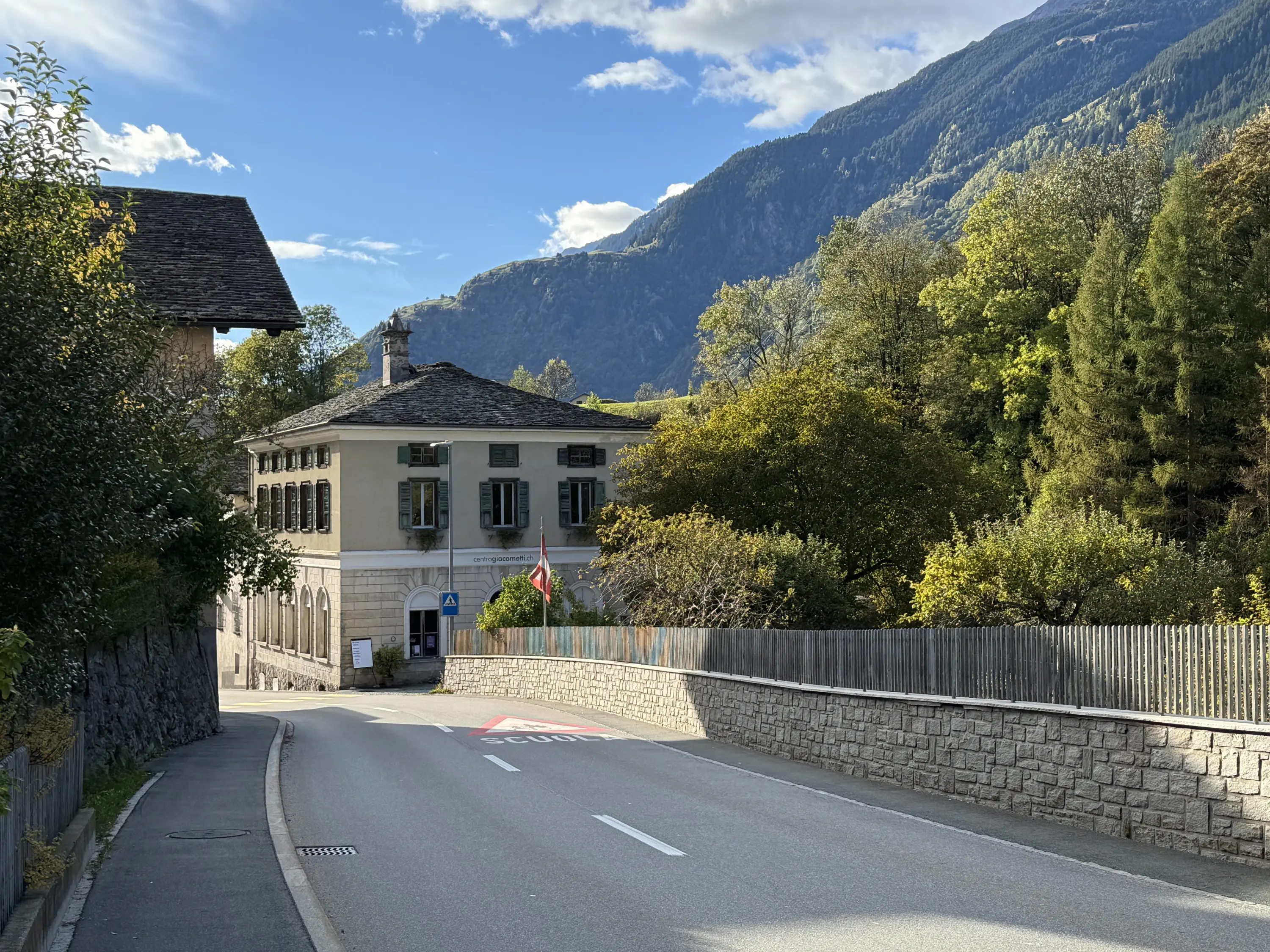 Un paesaggio montano con una strada che conduce a una casa storica con finestre verdi. Alle spalle, una montagna maestosa e alberi verdi. Un cielo sereno con alcune nuvole.