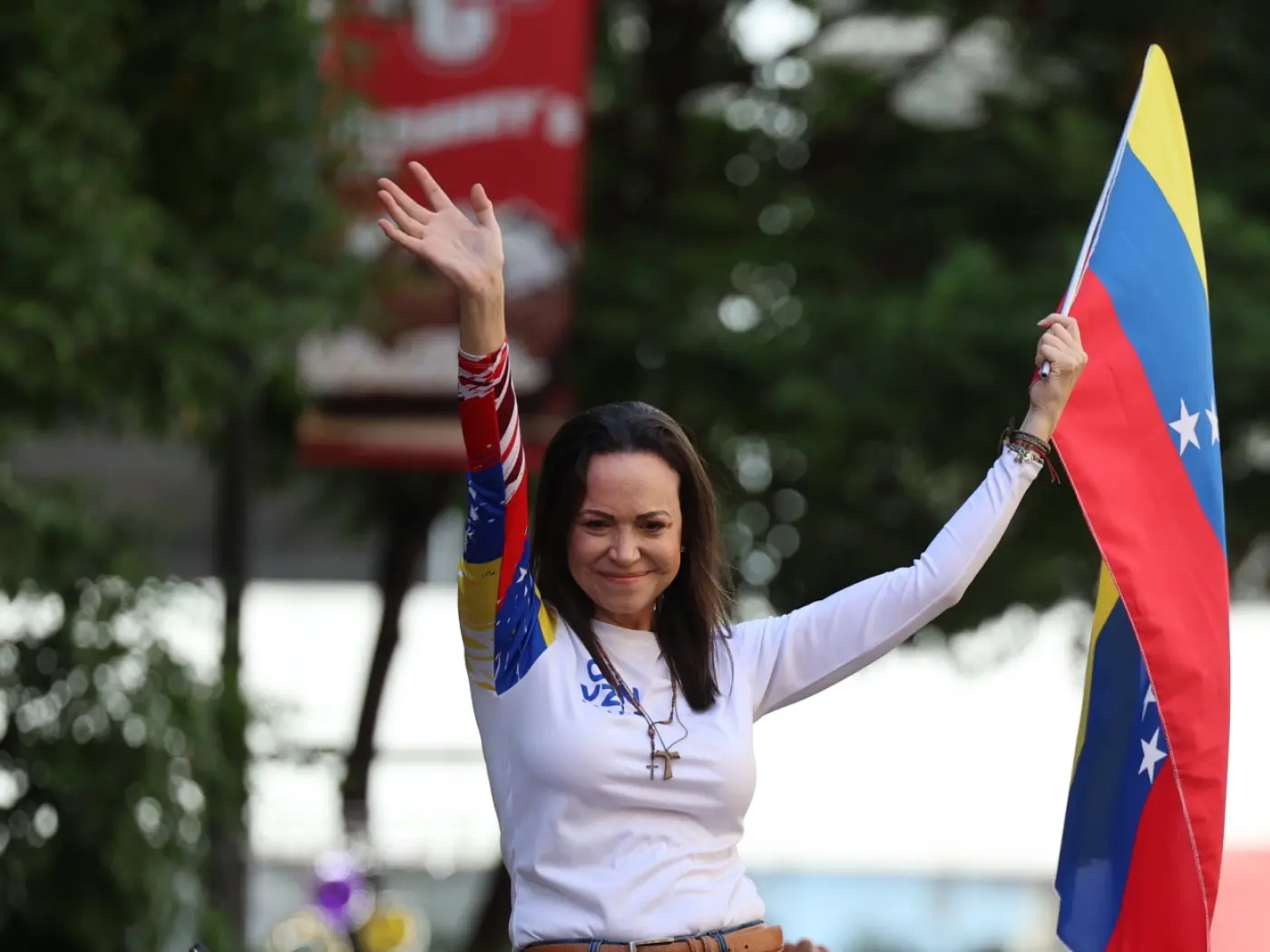 Donna sorridente con un sorriso amichevole, mentre saluta il pubblico e regge una bandiera venezuelana. È vestita con una maglietta bianca e appare in un contesto festoso, con piante verdi sullo sfondo.