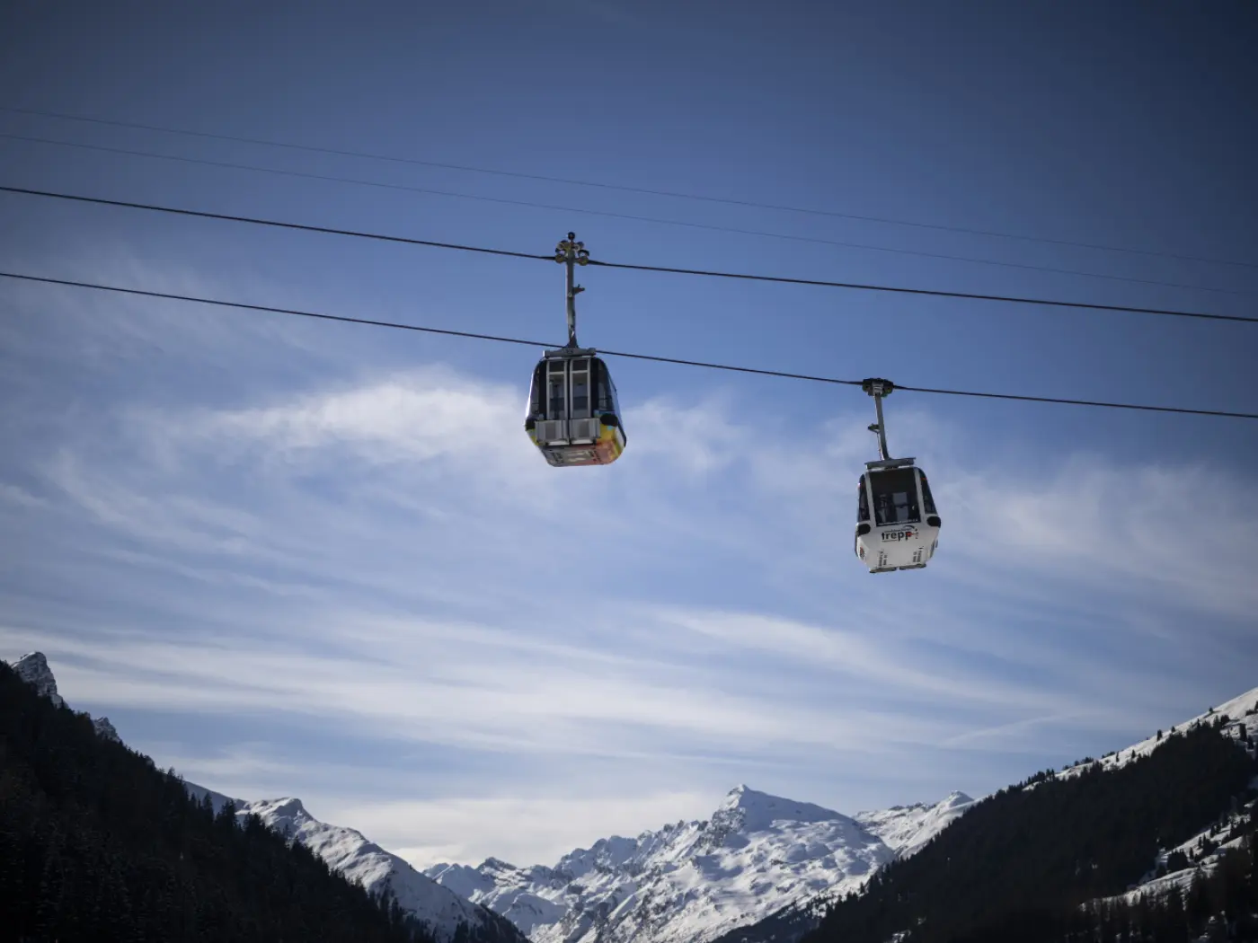 Due funivie che si muovono lungo un cavo sopra un paesaggio montano innevato, sotto un cielo azzurro con nuvole leggere.