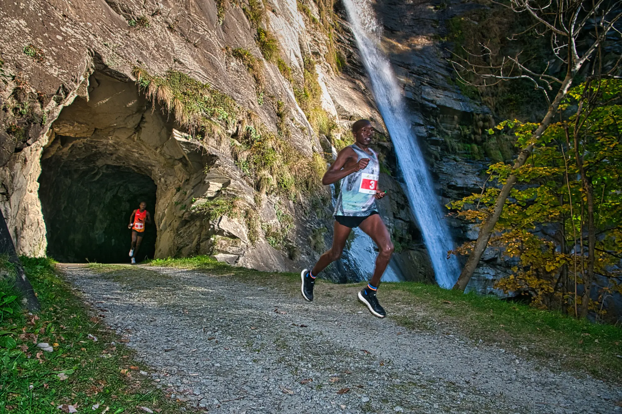 Due corridori competono lungo un sentiero di montagna. Una cascata scorre a destra e un tunnel di roccia si trova a sinistra. Un corridore indossa una maglietta bianca con il numero 13, mentre l'altro è in una maglietta rossa.
