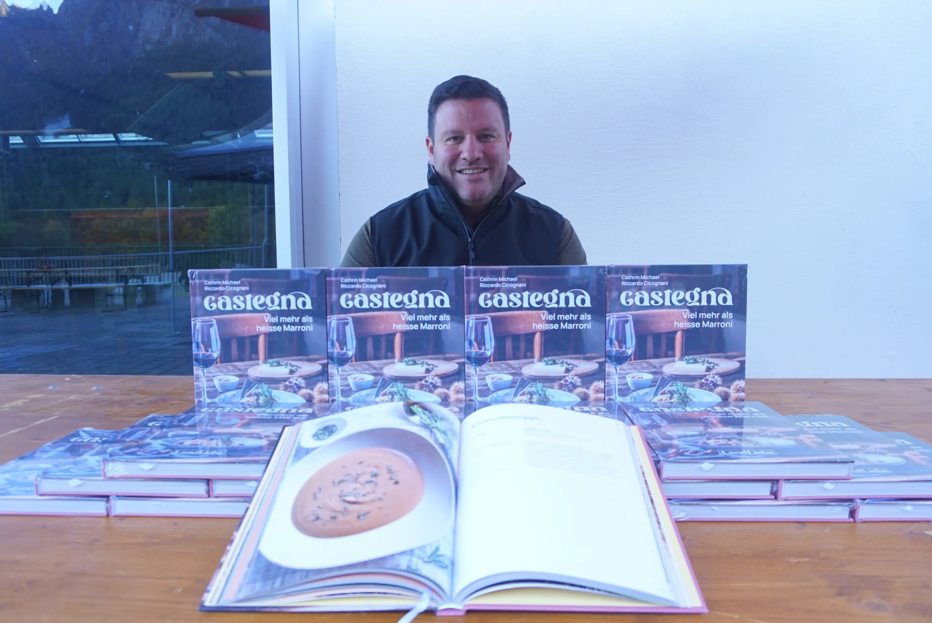 Un uomo sorridente è seduto dietro a una tavola con diversi libri di cucina aperti e impilati. Uno dei libri mostra una ricetta per una crema di funghi. Sullo sfondo si vedono paesaggi naturali.