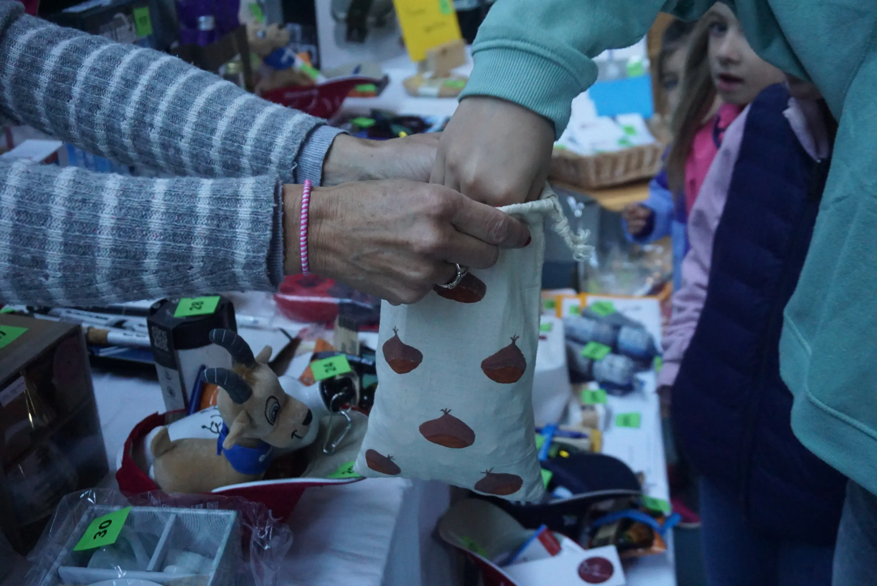 Due mani si scambiano un sacchetto di stoffa con disegni di ghiande in un mercatino, mentre sullo sfondo si vedono vari oggetti in vendita e un bambino che osserva.