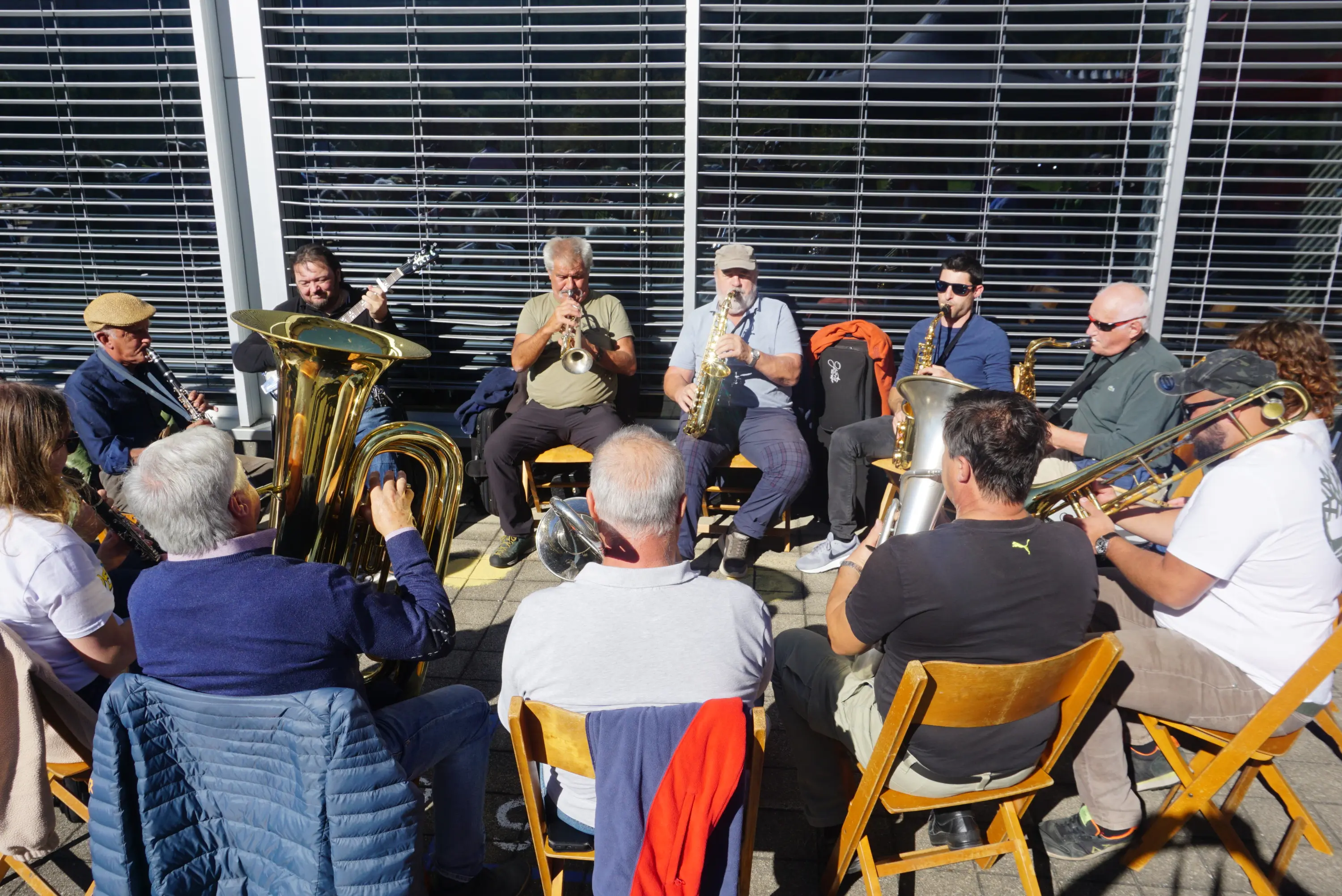 Gruppo di musicisti seduti su seggi chairs in un cerchio, mentre suonano strumenti a fiato e a percussione. Sono vestiti in modo casual, alcuni indossano cappelli. Sullo sfondo, ci sono persiane chiuse che filtrano la luce.