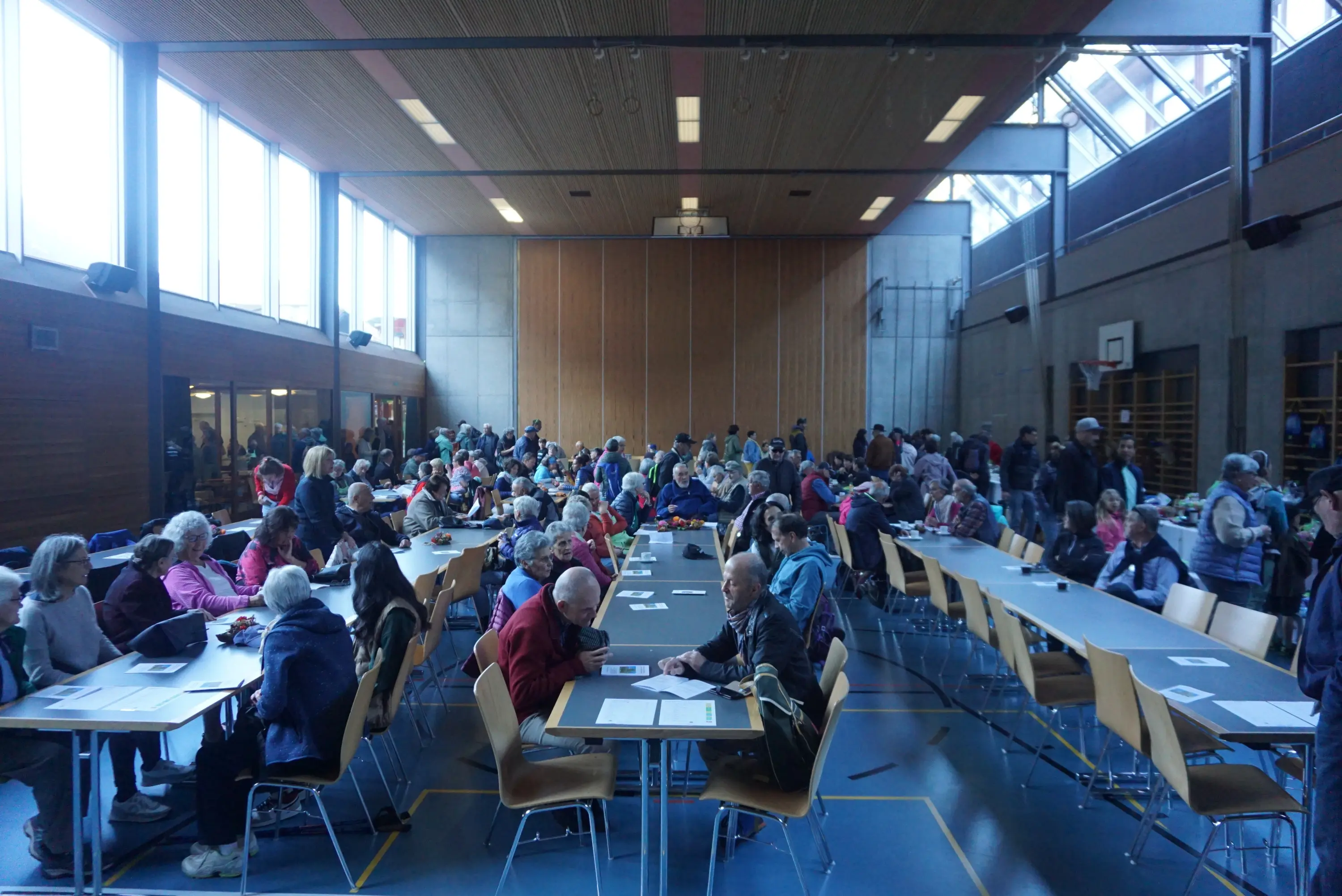 Una sala ampia e luminosa piena di persone anziane sedute attorno a tavoli. Molti partecipanti chiacchierano tra loro mentre alcuni sono concentrati su fogli di carta. La grande finestra lascia entrare abbondante luce naturale.