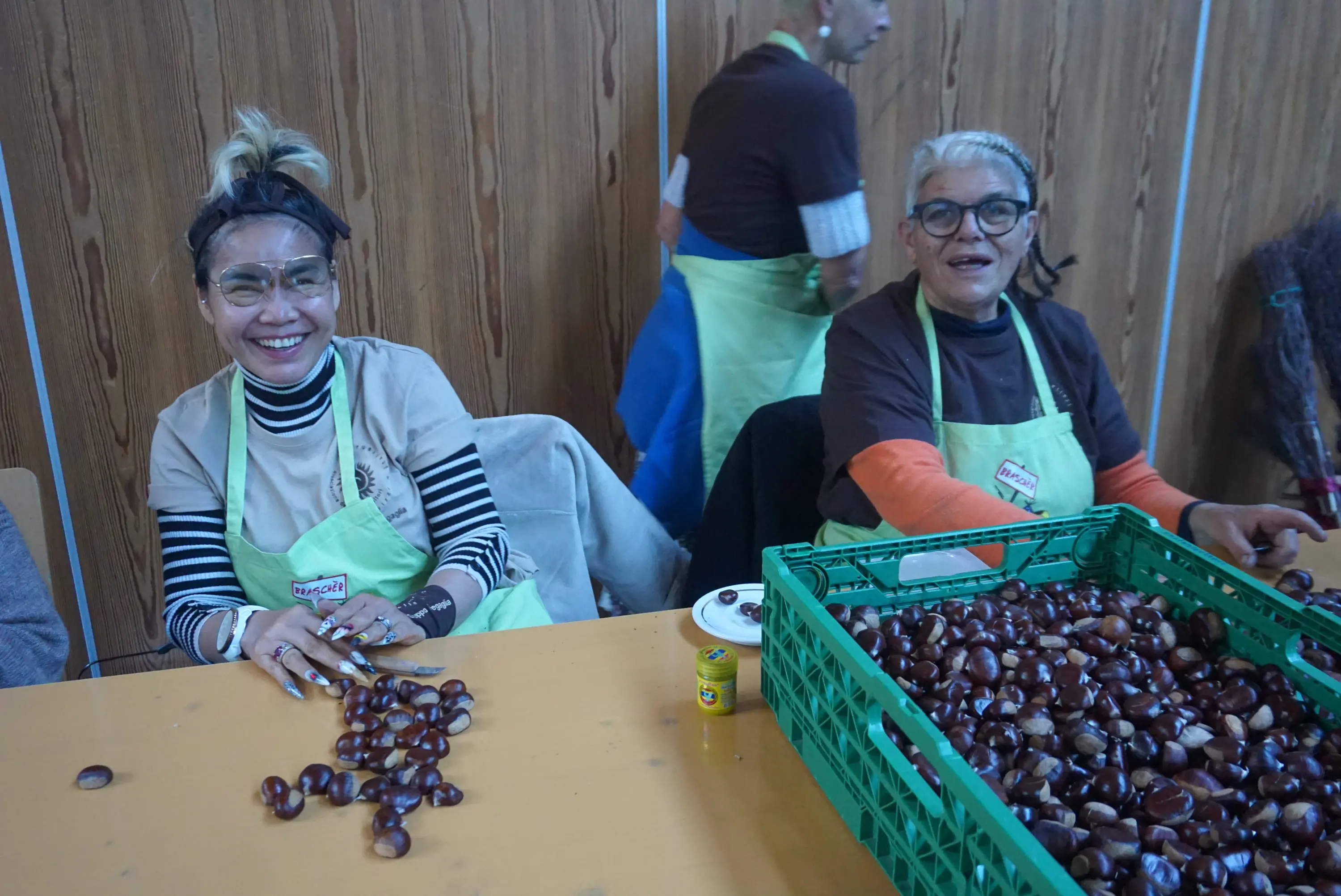 Due donne sorridenti sono sedute a un tavolo, mentre selezionano castagne. Indossano grembiuli verdi e occhiali protettivi. Dietro di loro, si vedono altre persone impegnate.