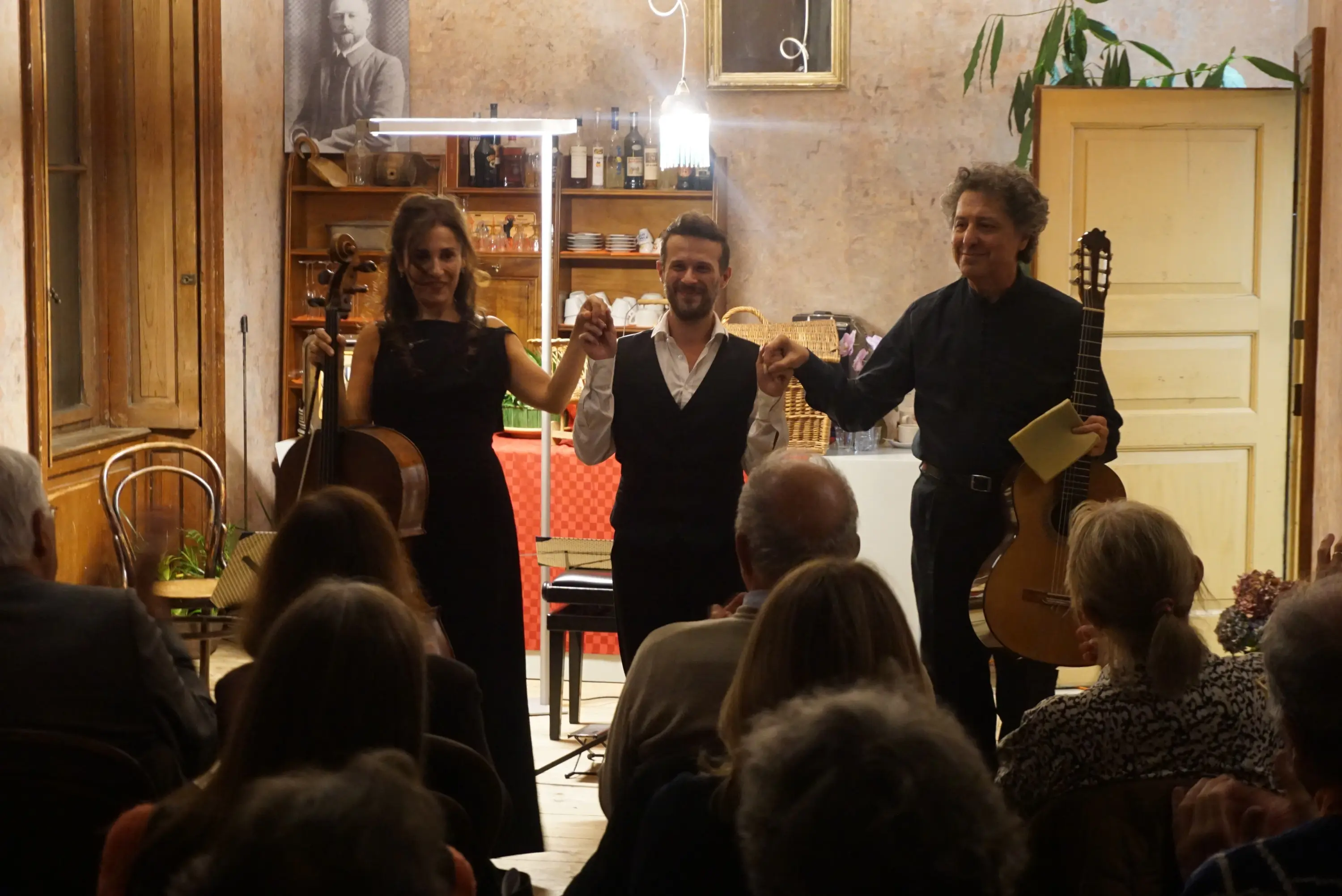 Tre musicisti applauditi sul palco, sorridenti e in piedi con le mani alzate. Due di loro tengono strumenti musicali: una violoncellista con il suo violoncello e un chitarrista con la chitarra. Il pubblico di spalle è visibile, immerso nell'atmosfera di un concerto intimo.